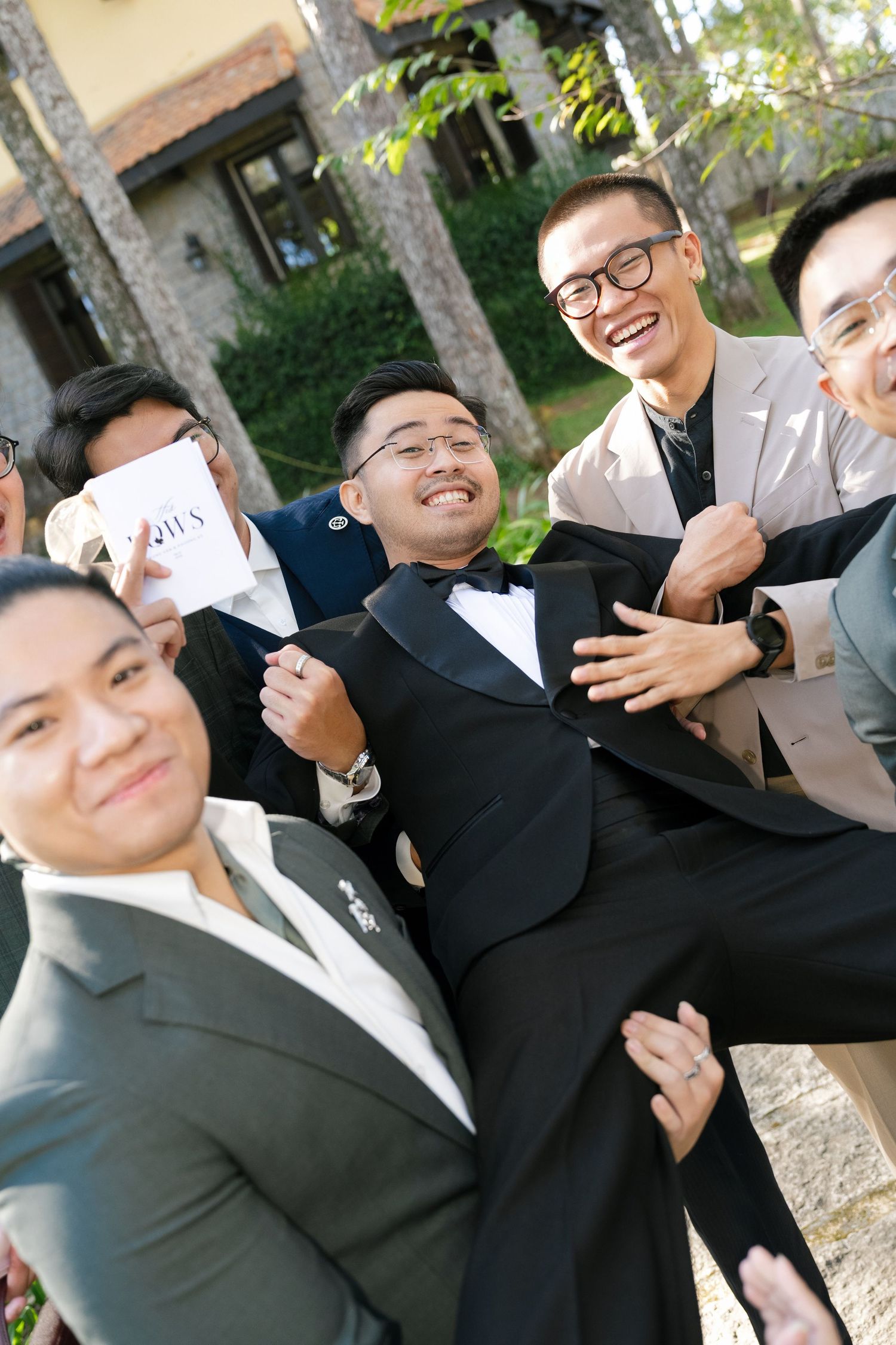 Laughing groomsmen lifting the groom in a black tuxedo outdoors at a wedding venue.
