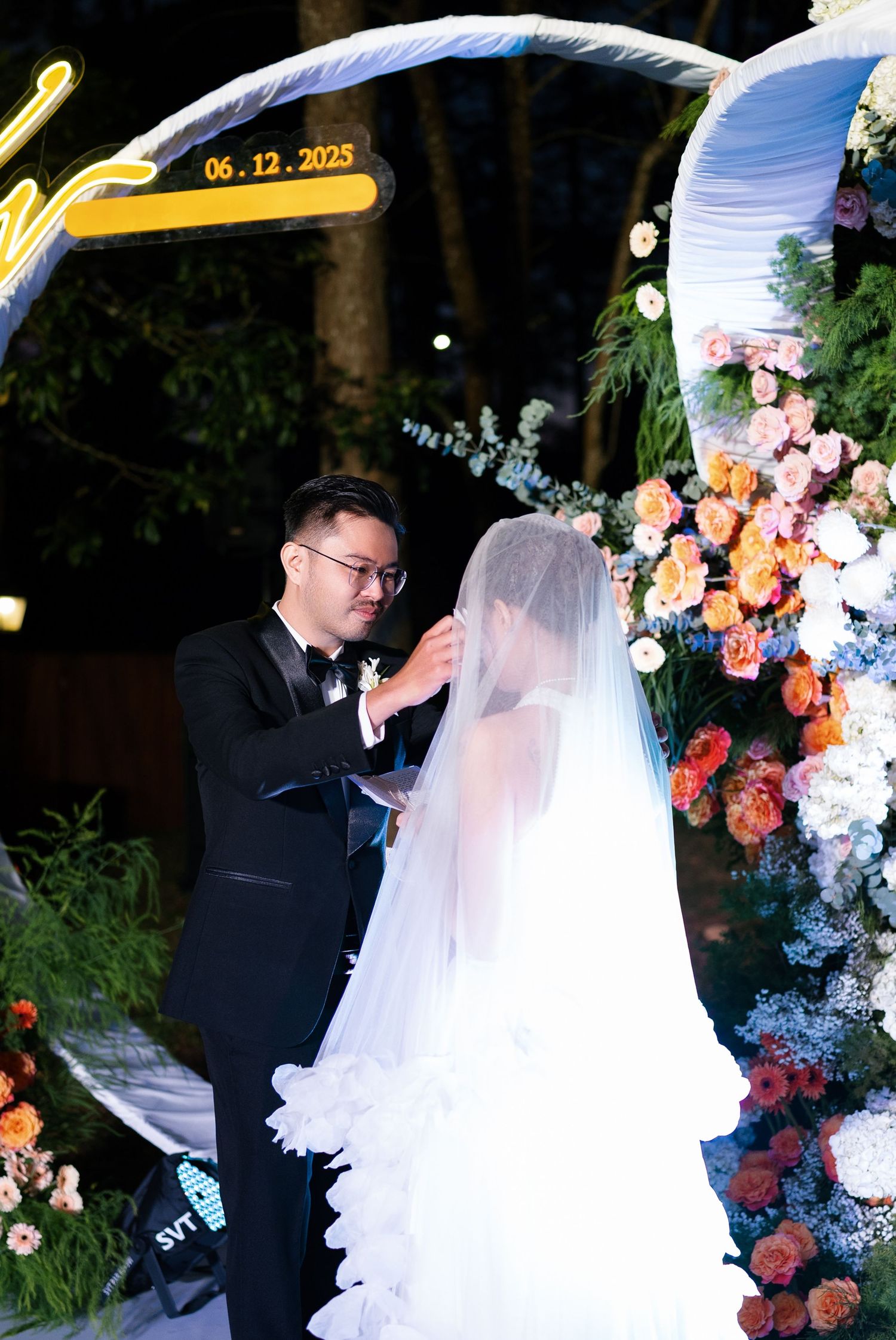 Groom lifts bride's veil during outdoor wedding ceremony under floral arch on December 6, 2025.