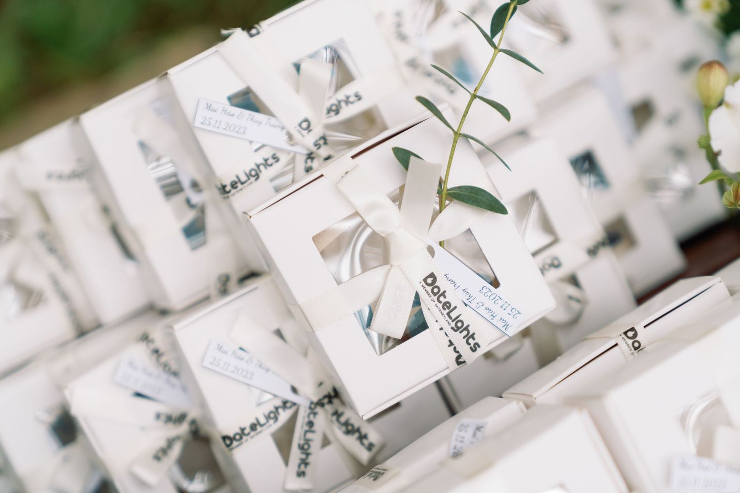 White wedding favor boxes tied with silver ribbons and decorated with greenery, arranged in rows at an outdoor event.