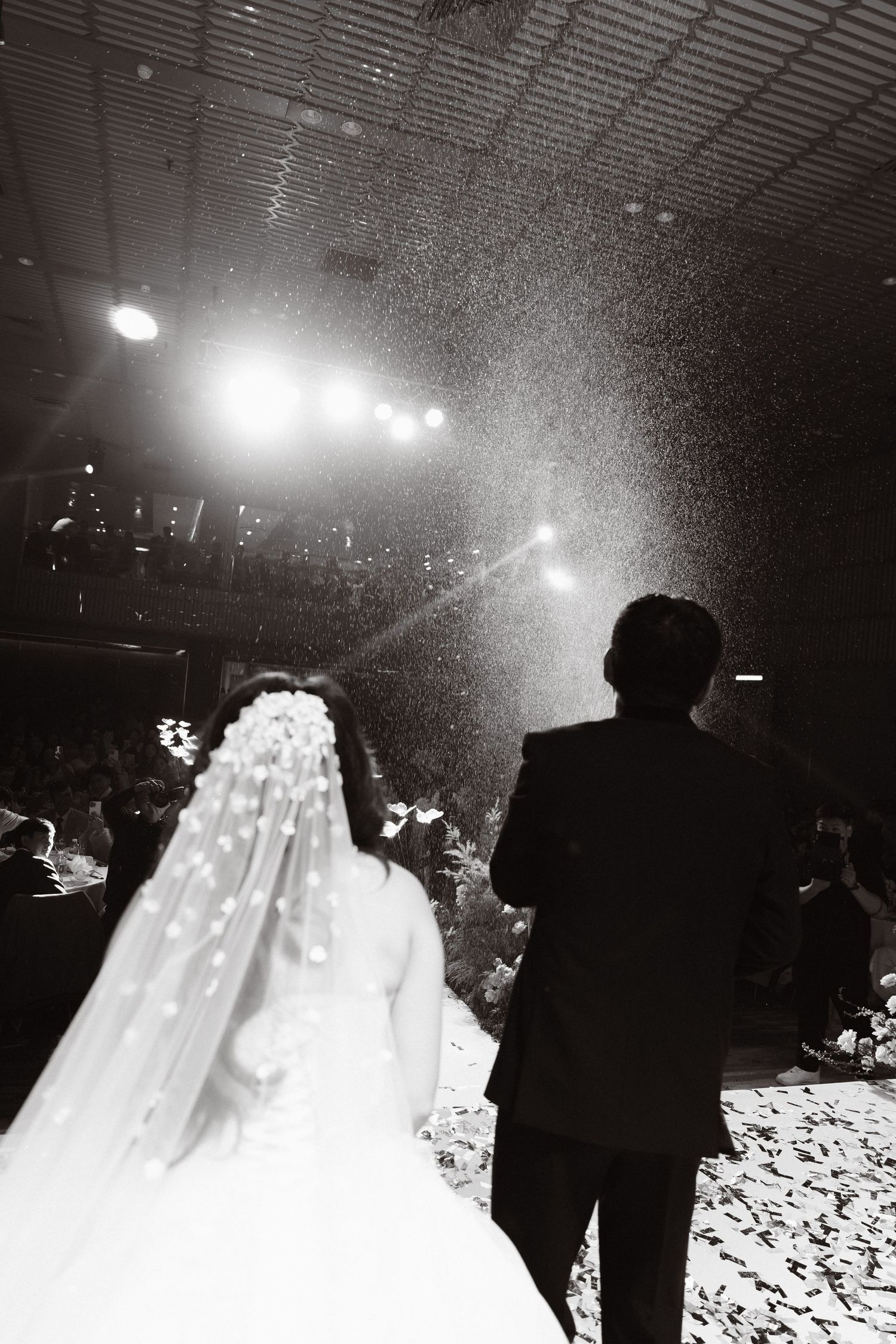 Black and white photo of a bride and groom walking down the aisle with confetti falling at their wedding reception.
