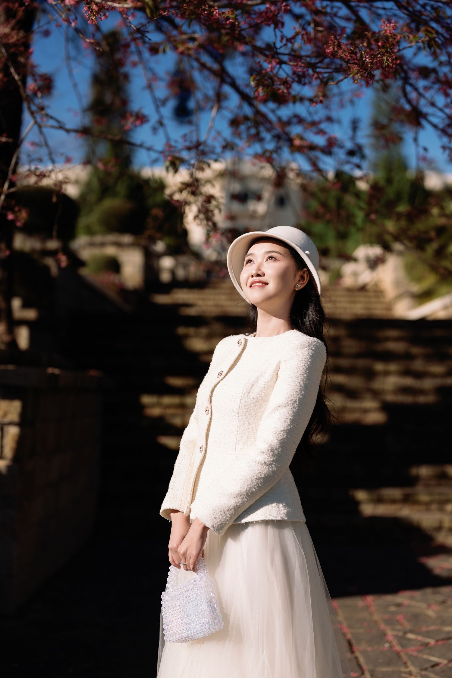 Young woman in white tweed jacket, skirt, and bucket hat holding a pearl bag under cherry blossom tree.