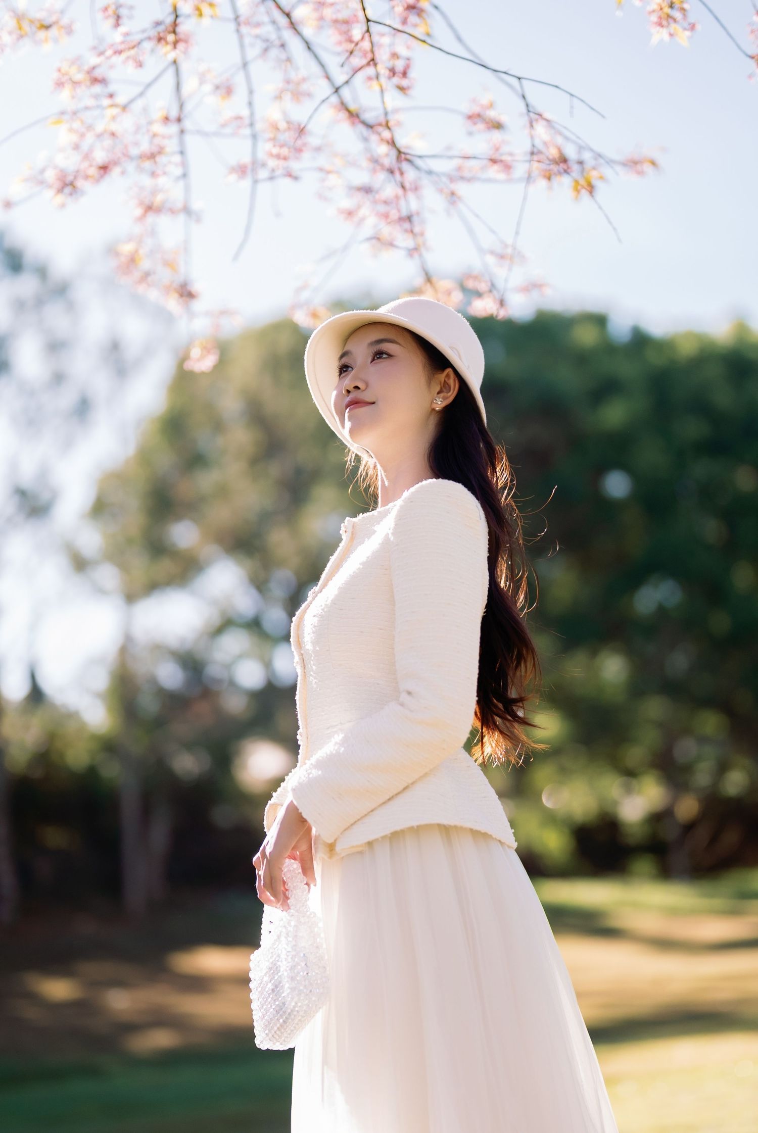 Young woman in white dress and hat stands gracefully under blooming cherry blossoms in a sunlit park.