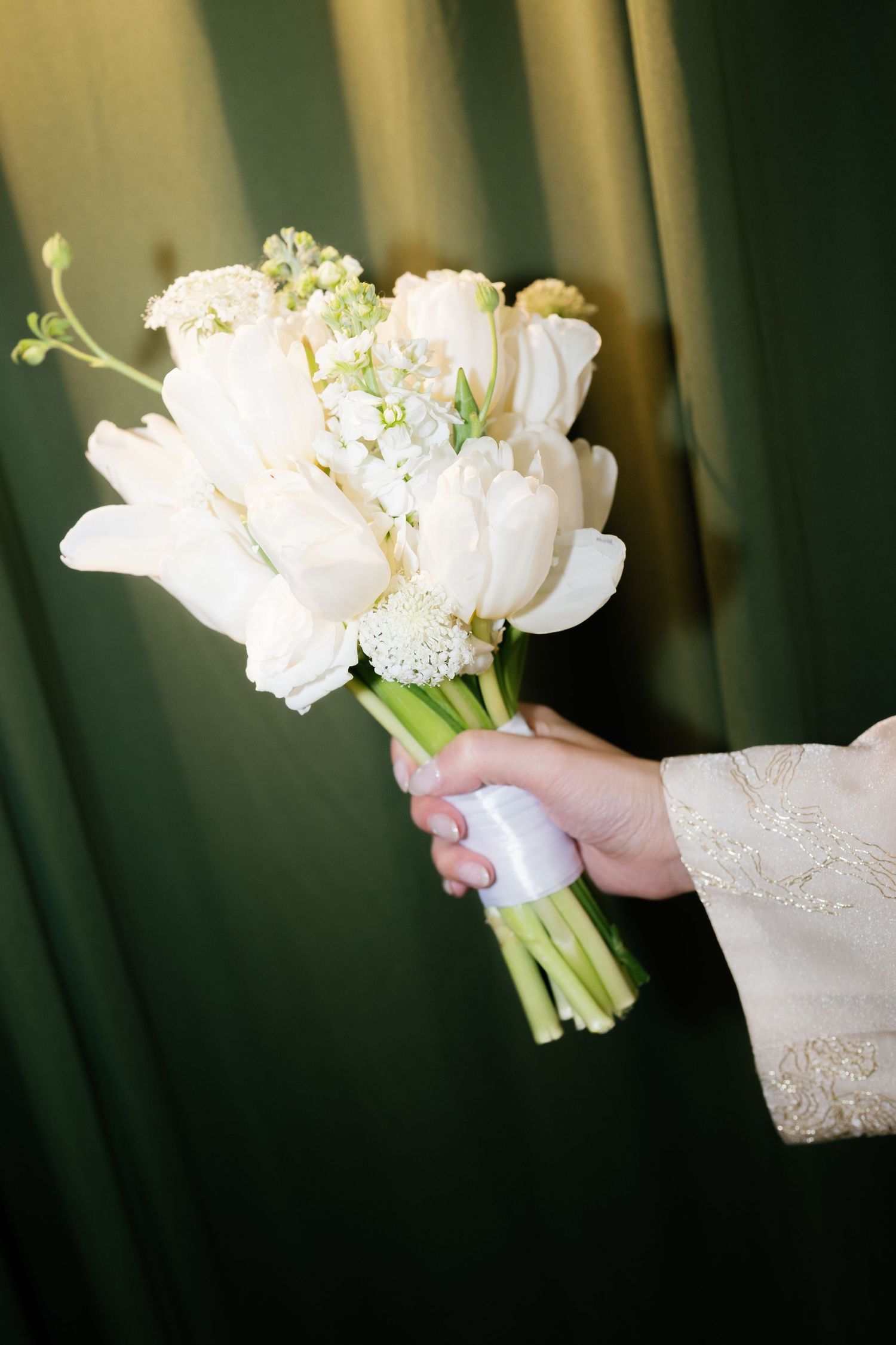 Hand holding a white tulip bridal bouquet with ribbon wrap against a green curtain backdrop.