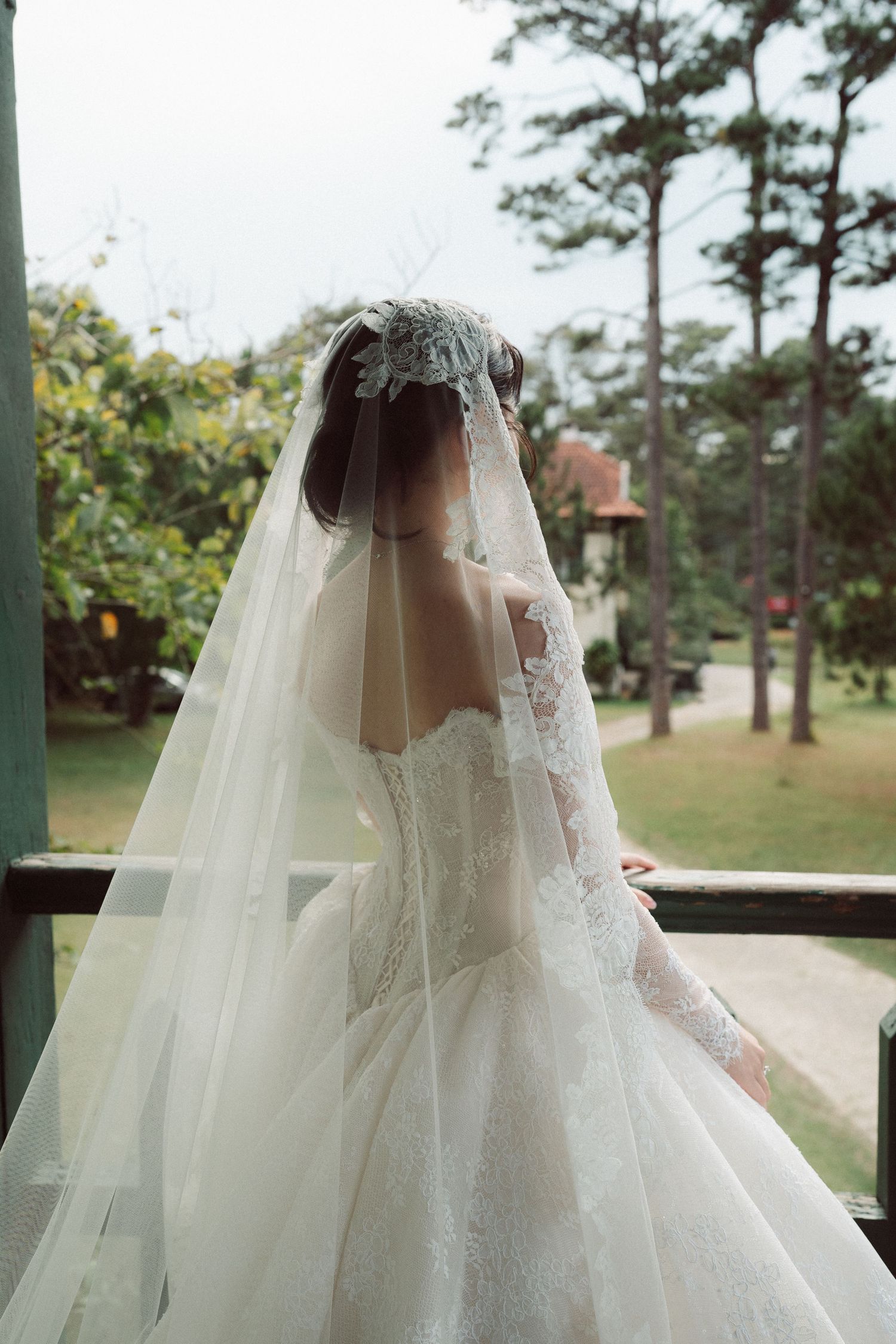 Bride in elegant lace wedding gown with long veil and floral headpiece, standing on balcony overlooking lush garden.