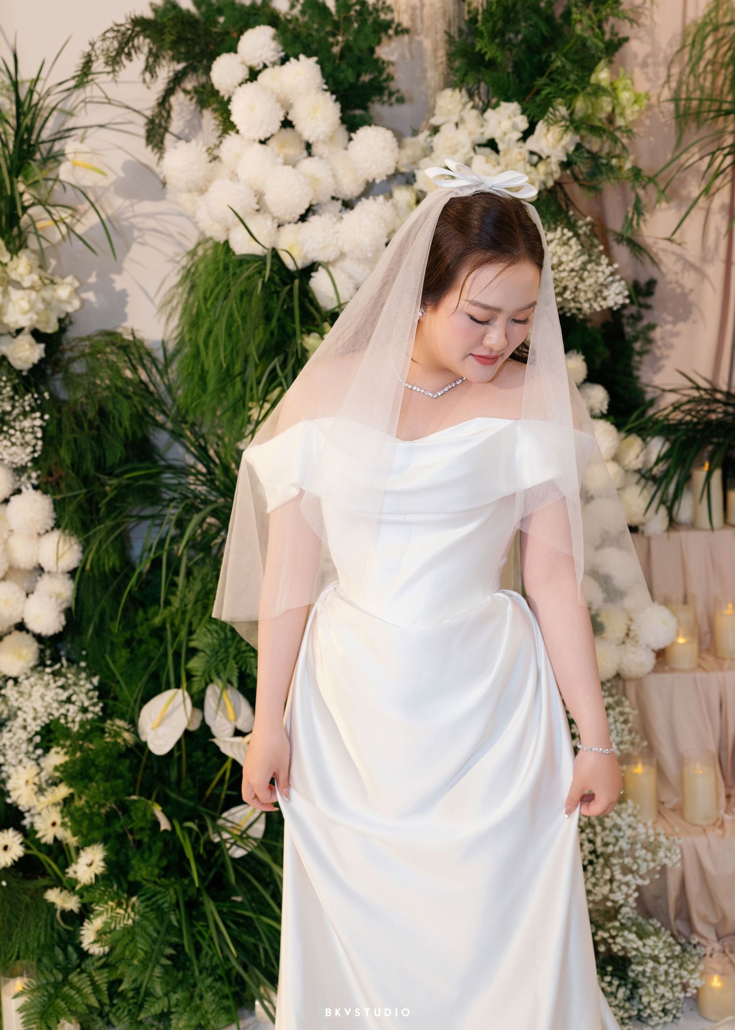 A bride in a white satin off-shoulder gown and veil poses before a lush floral backdrop of white blooms and greenery.