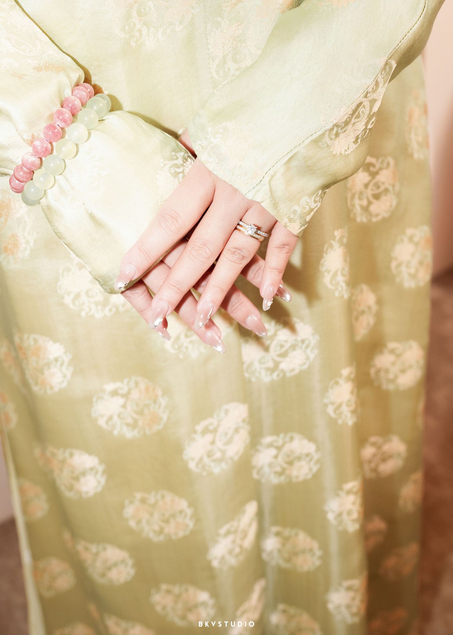 Close-up of hands with silver rings and pink beaded bracelet, wearing a light green floral embroidered traditional outfit.