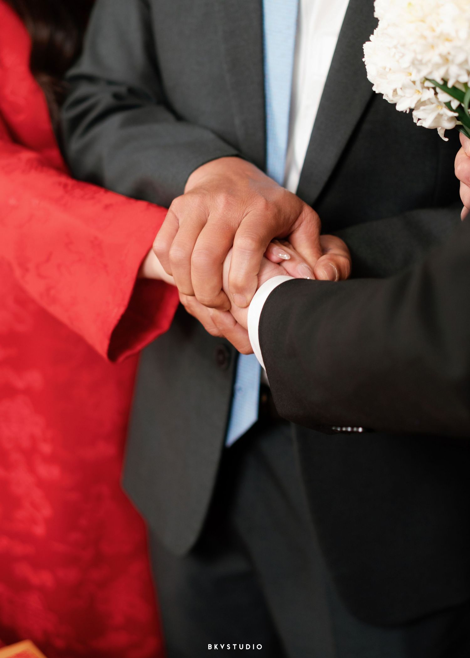 Close-up of a couple holding hands during a wedding ceremony, groom in grey suit holding white flowers.