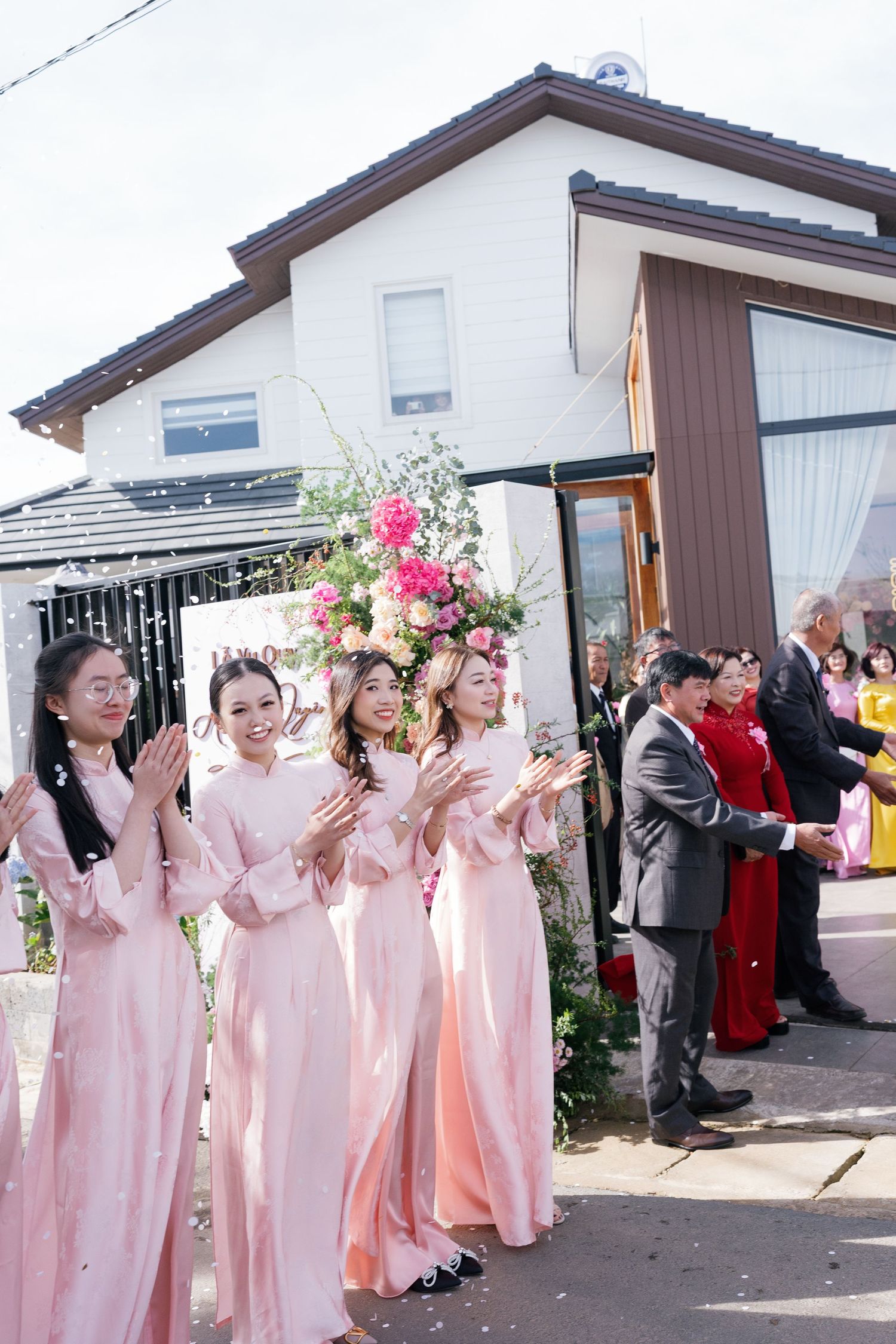 Bridesmaids in pink ao dai dresses clapping at a Vietnamese wedding ceremony outdoors.