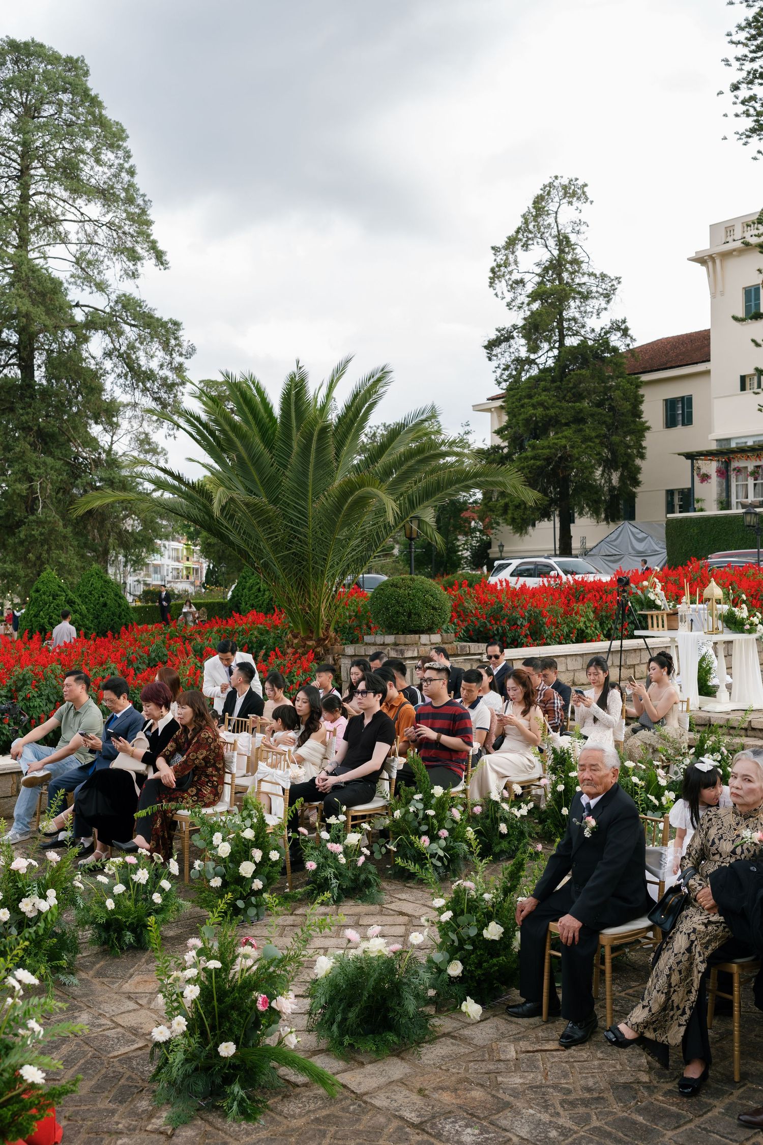 Outdoor wedding ceremony with guests seated on gold chairs amid white floral arrangements, palm trees, and red flowers in a garden.