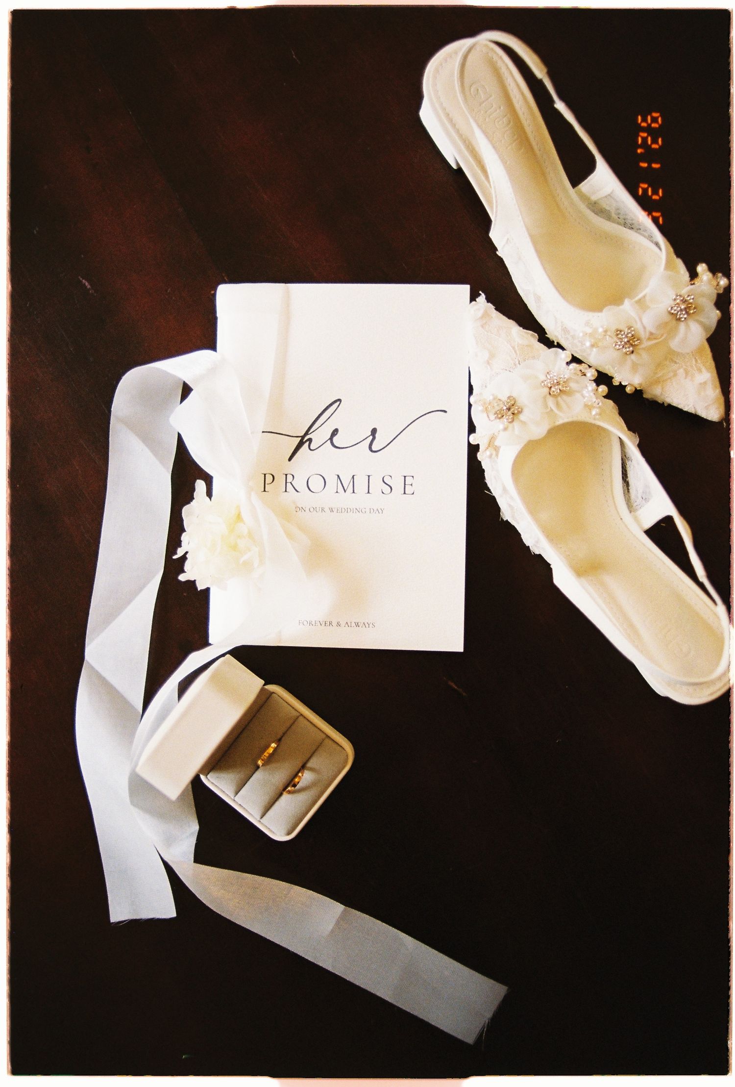 Elegant bridal flat shoes, Her Promise vow book, and gold wedding rings in a velvet box on dark wood surface.