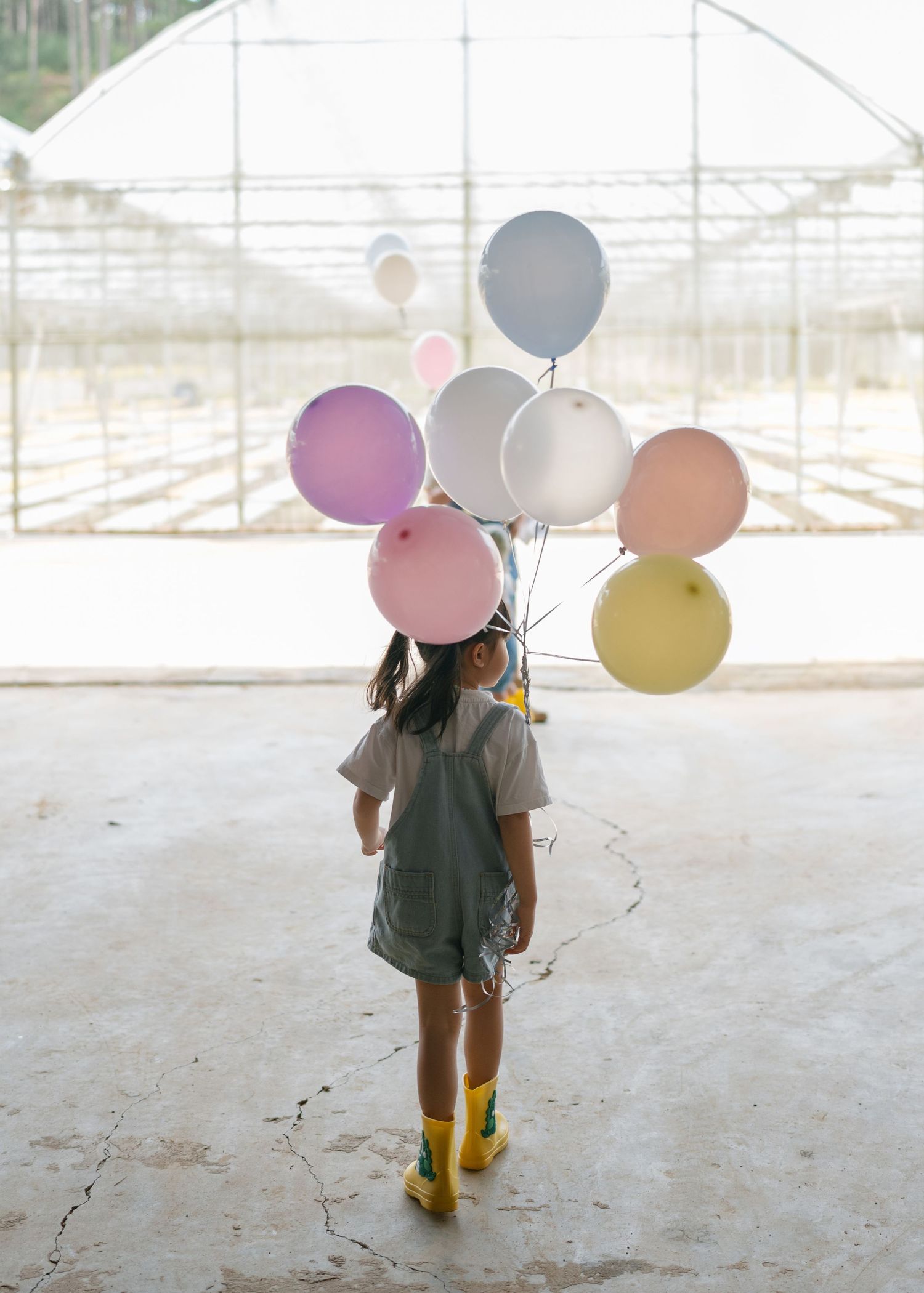 A young girl in overalls and yellow boots holds colorful pastel balloons inside a bright greenhouse.