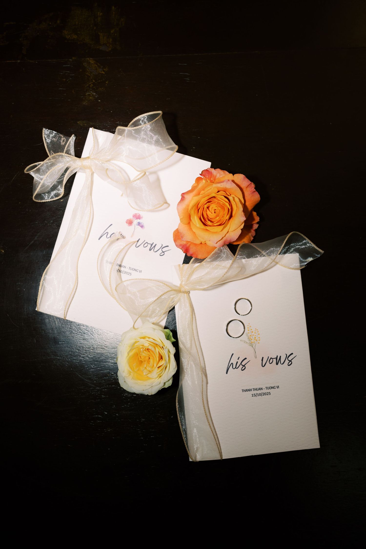 Two elegant wedding vow booklets tied with sheer ribbon, adorned with orange and yellow roses on a dark surface.