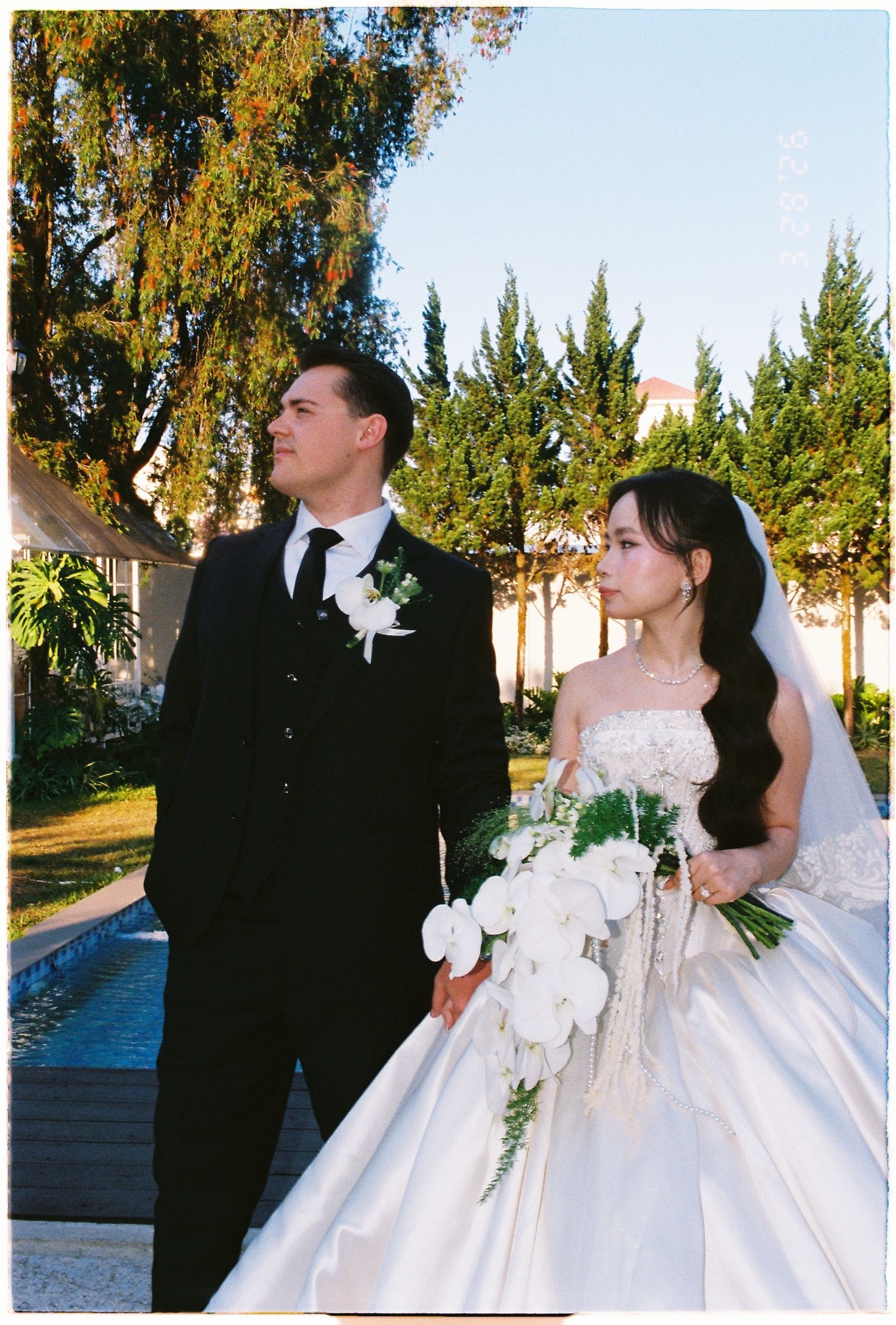 Bride in white gown and groom in black suit posing outdoors at a sunny wedding venue.