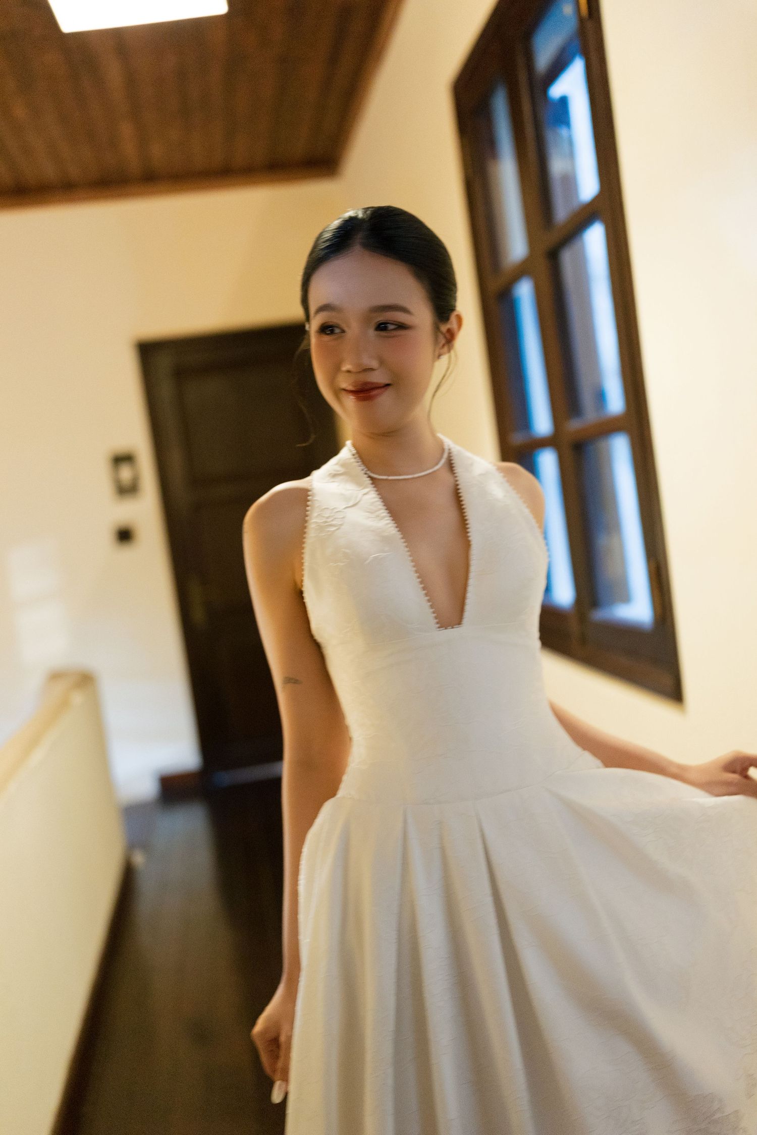 Elegant bride in white halter wedding gown with pearl necklace smiling in a warmly lit hallway.