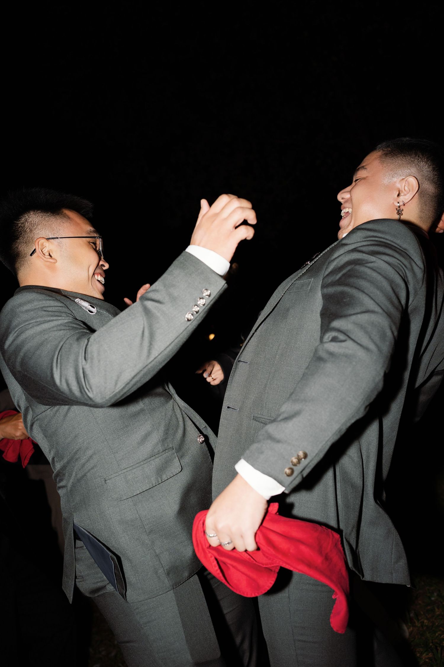 Two groomsmen in grey suits laughing and celebrating joyfully at outdoor evening wedding reception.