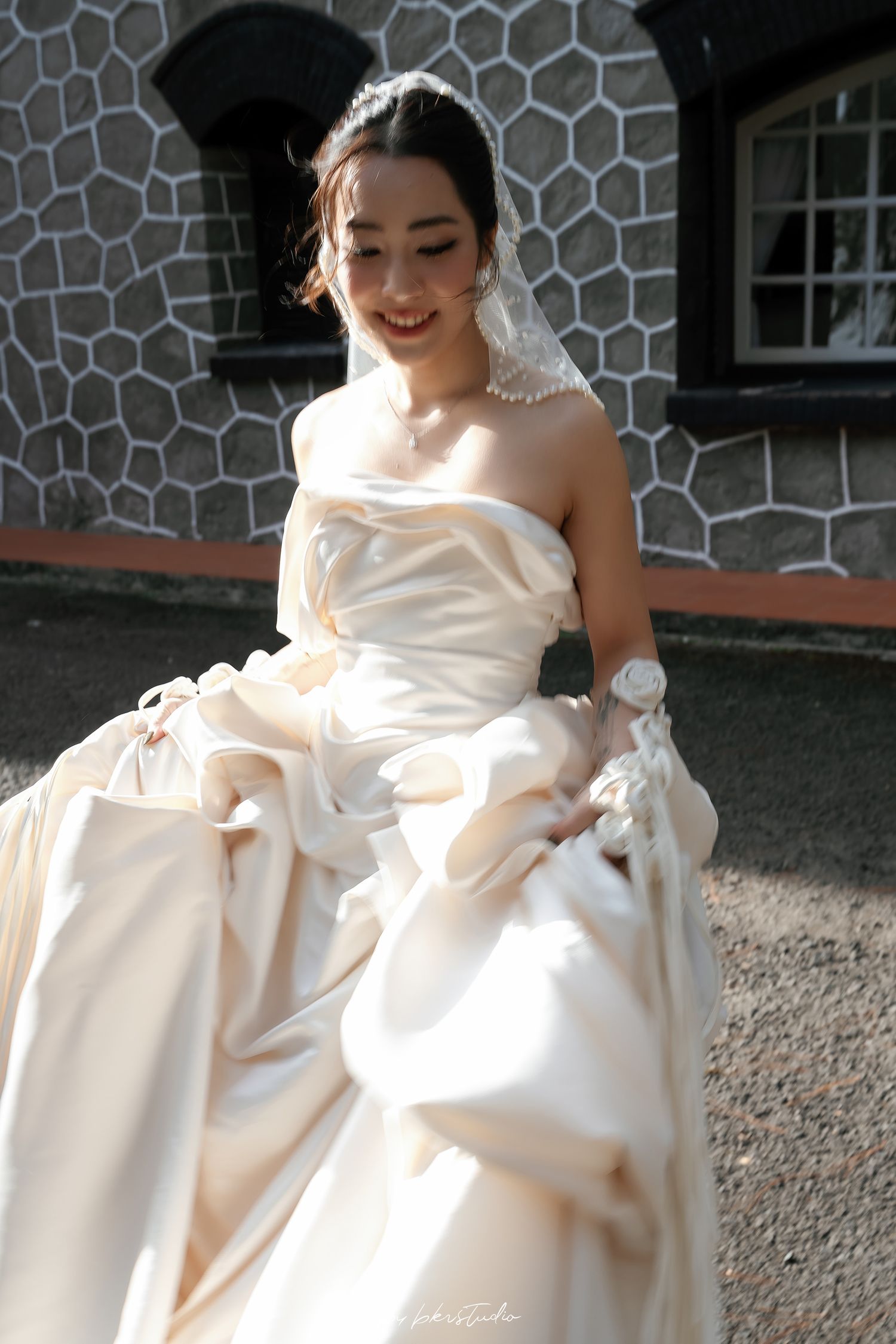 Smiling bride in strapless ivory satin wedding gown with veil and pearl necklace posing outdoors.