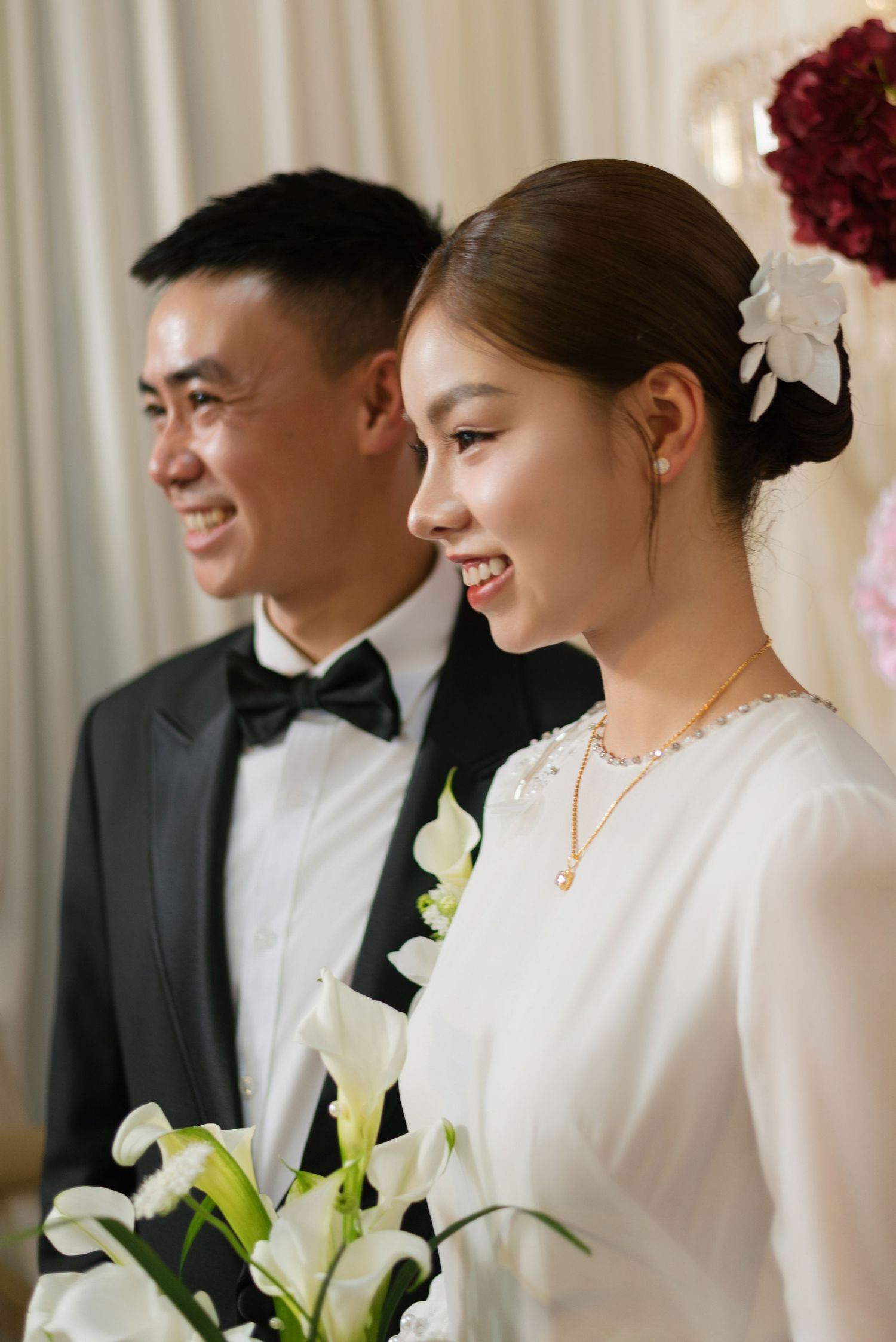 Smiling Asian bride in white dress holding calla lilies stands beside groom in black tuxedo at wedding ceremony.