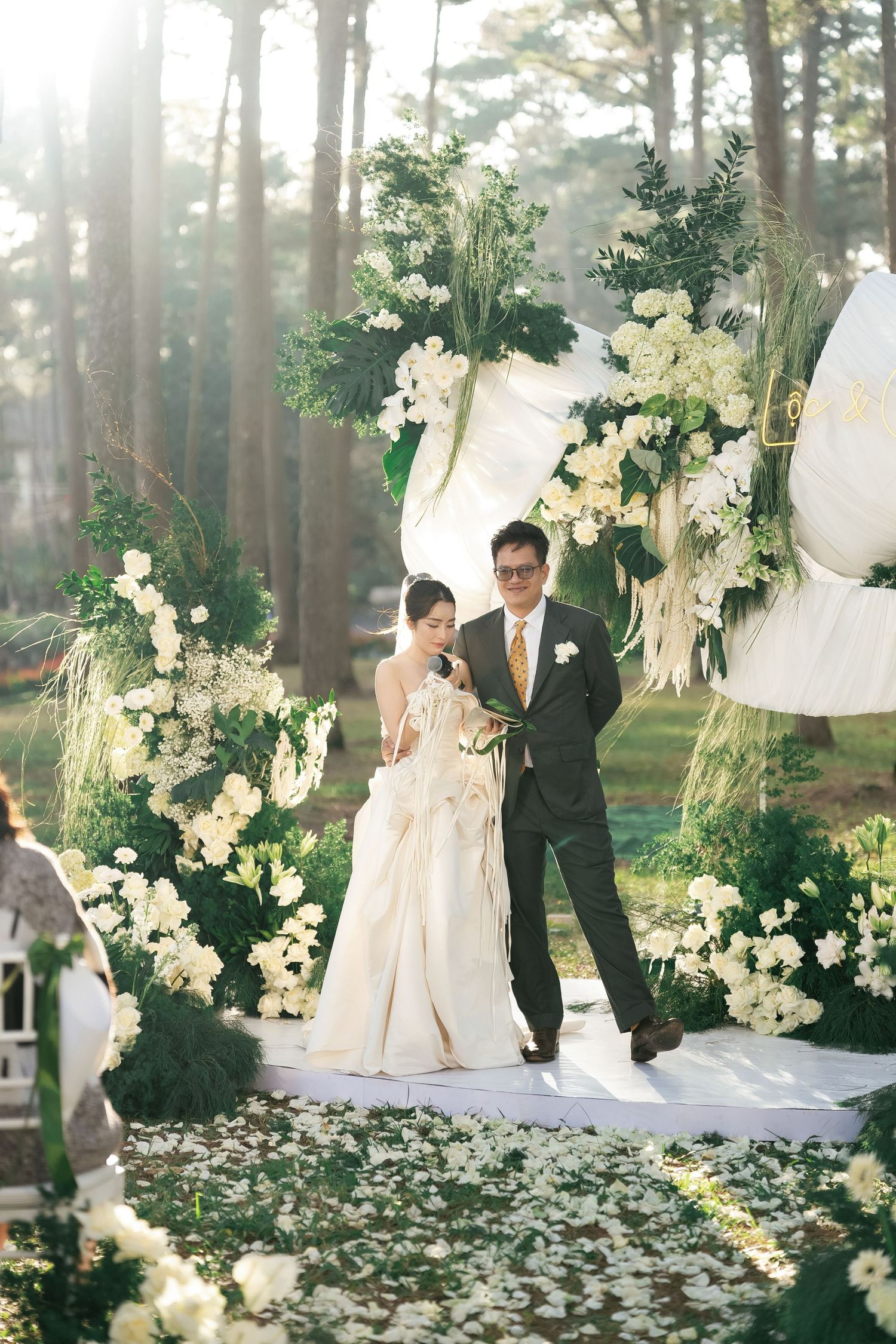 Bride and groom stand at an outdoor wedding altar adorned with white flowers and greenery in a sunlit forest setting.