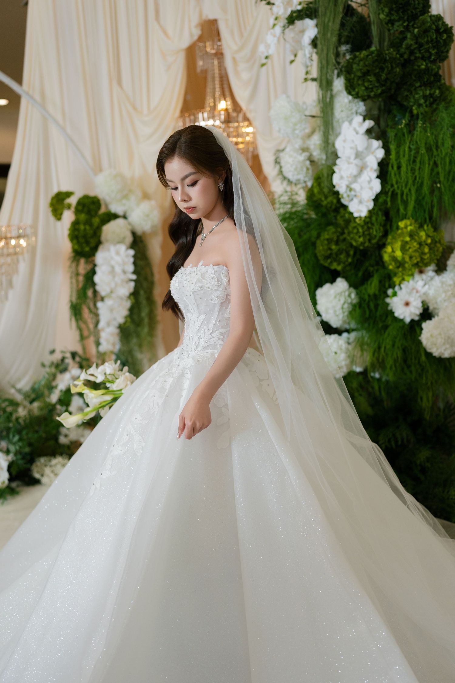 A young Asian bride in a white ballgown with lace bodice and veil poses among green and white floral wedding decorations.