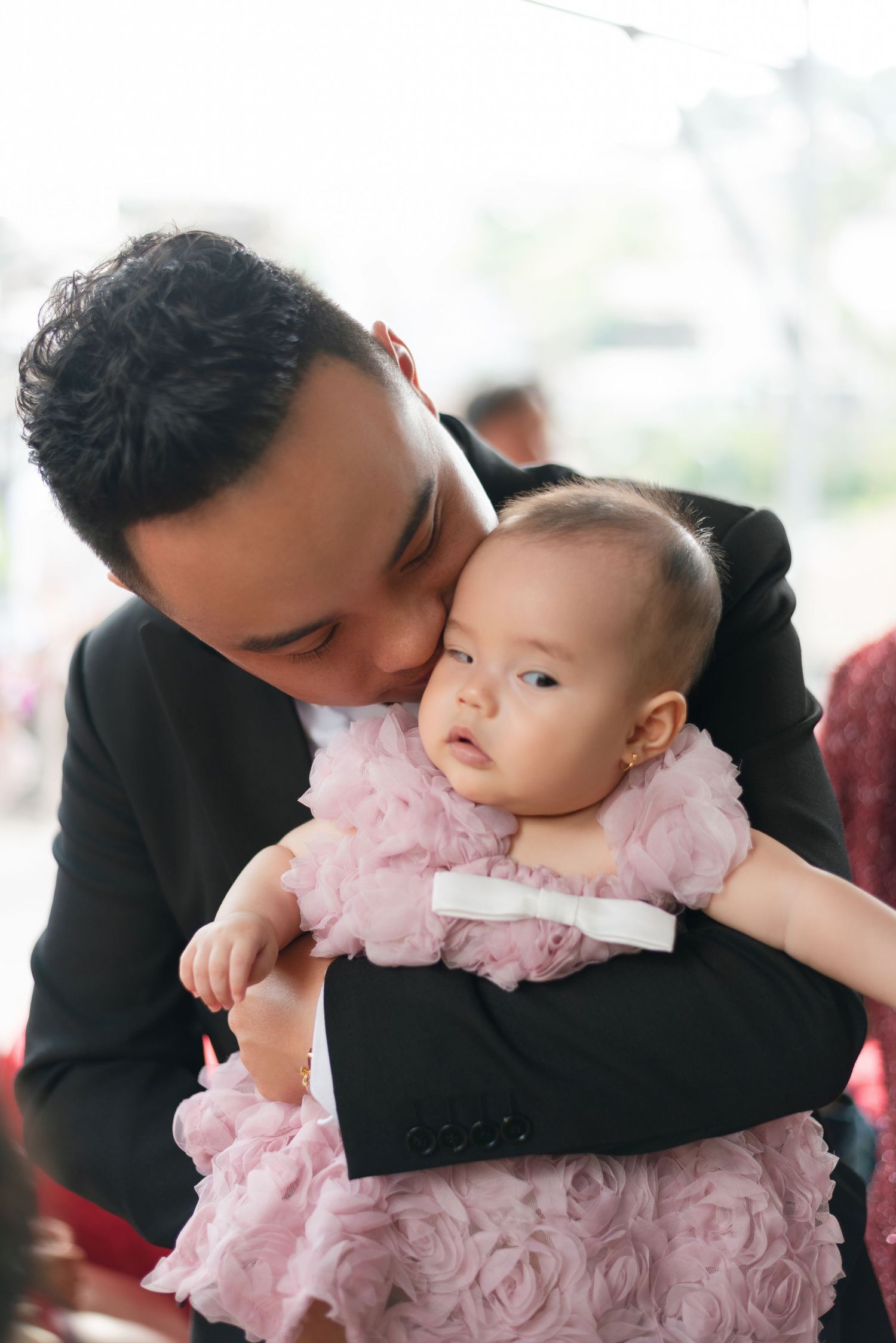 A man in a black suit tenderly kisses a baby girl in a pink ruffled dress while holding her gently.