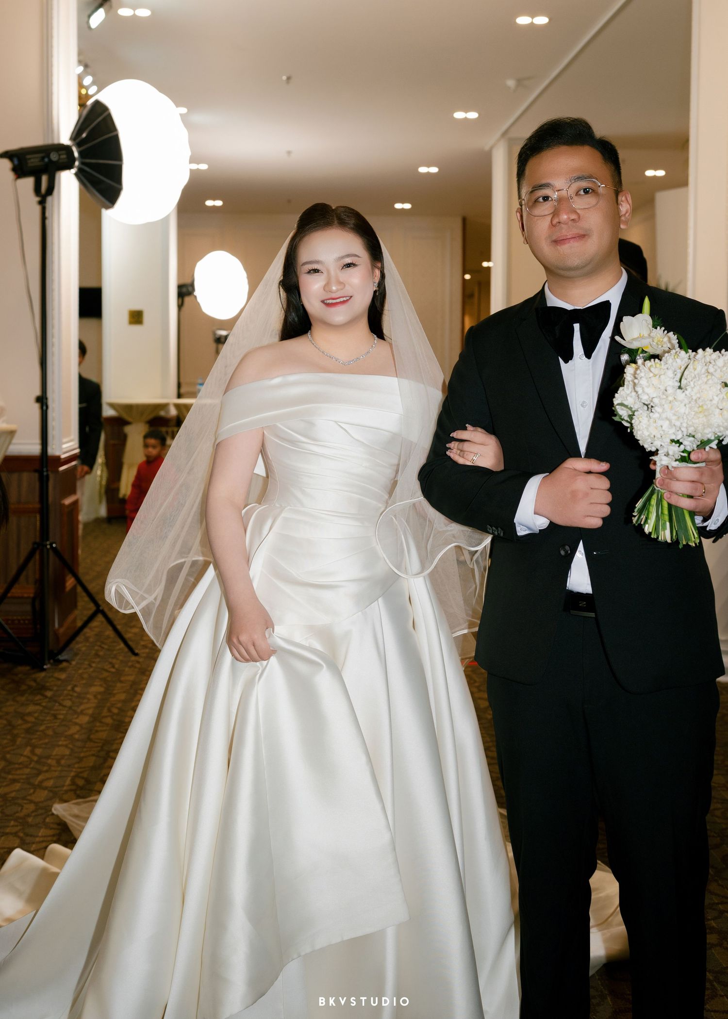 Bride in white off-shoulder satin ball gown with veil stands with groom in black tuxedo holding white bouquet.