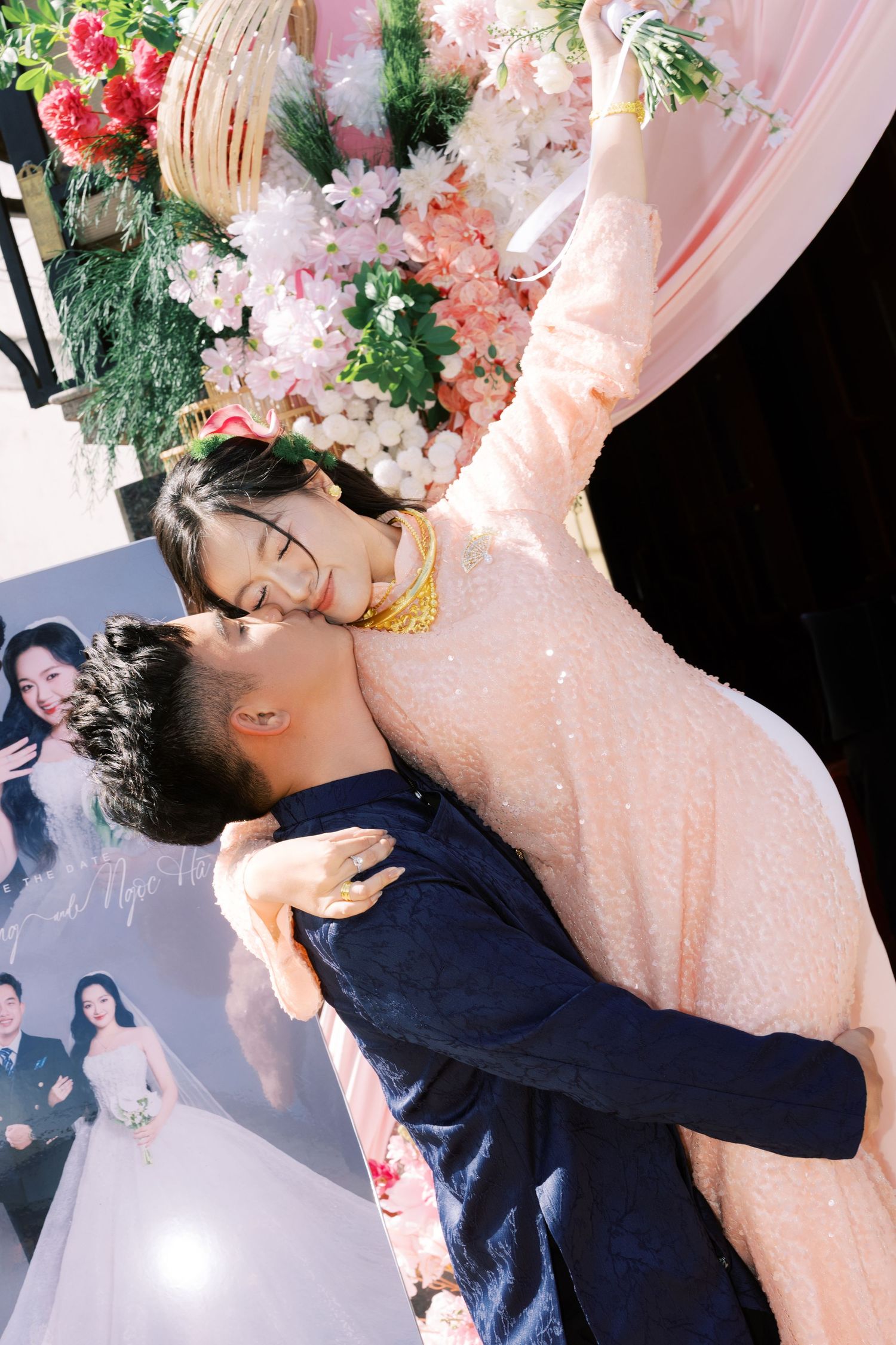 Couple sharing a romantic embrace at a wedding reception, woman in pink sequin dress with gold necklace, floral backdrop.