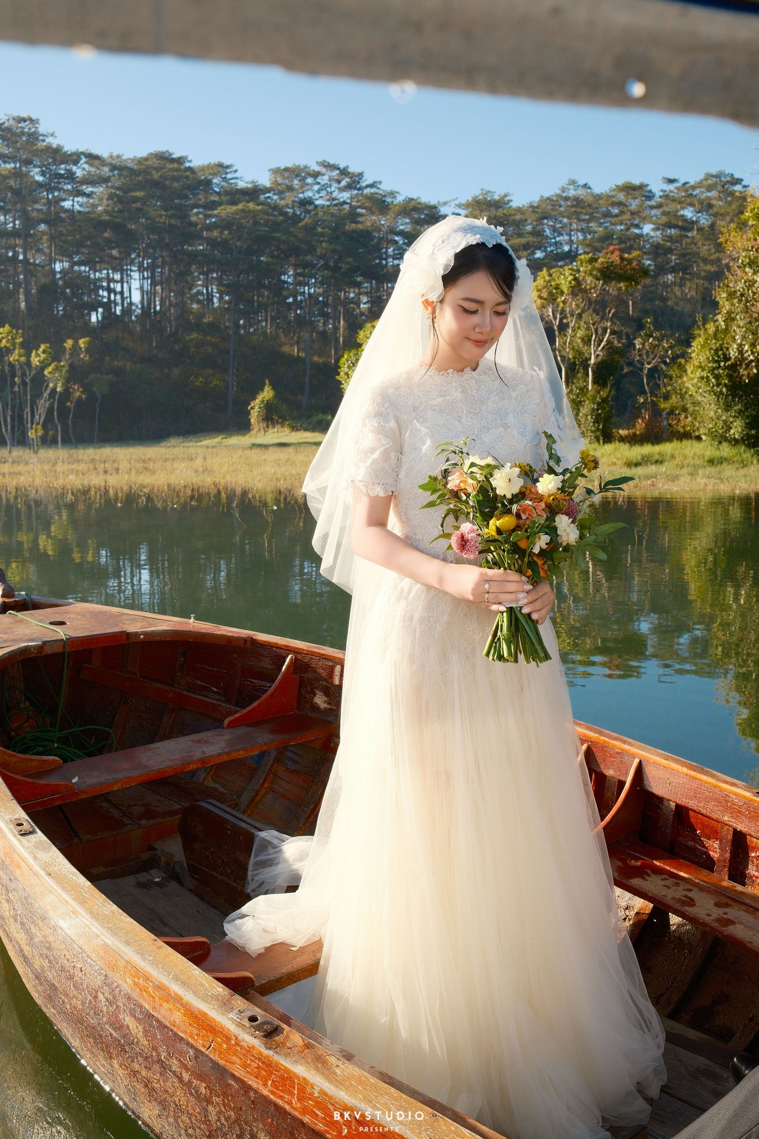 Elegant bride in lace gown holds a colorful bouquet while standing in a wooden boat on a serene lake.