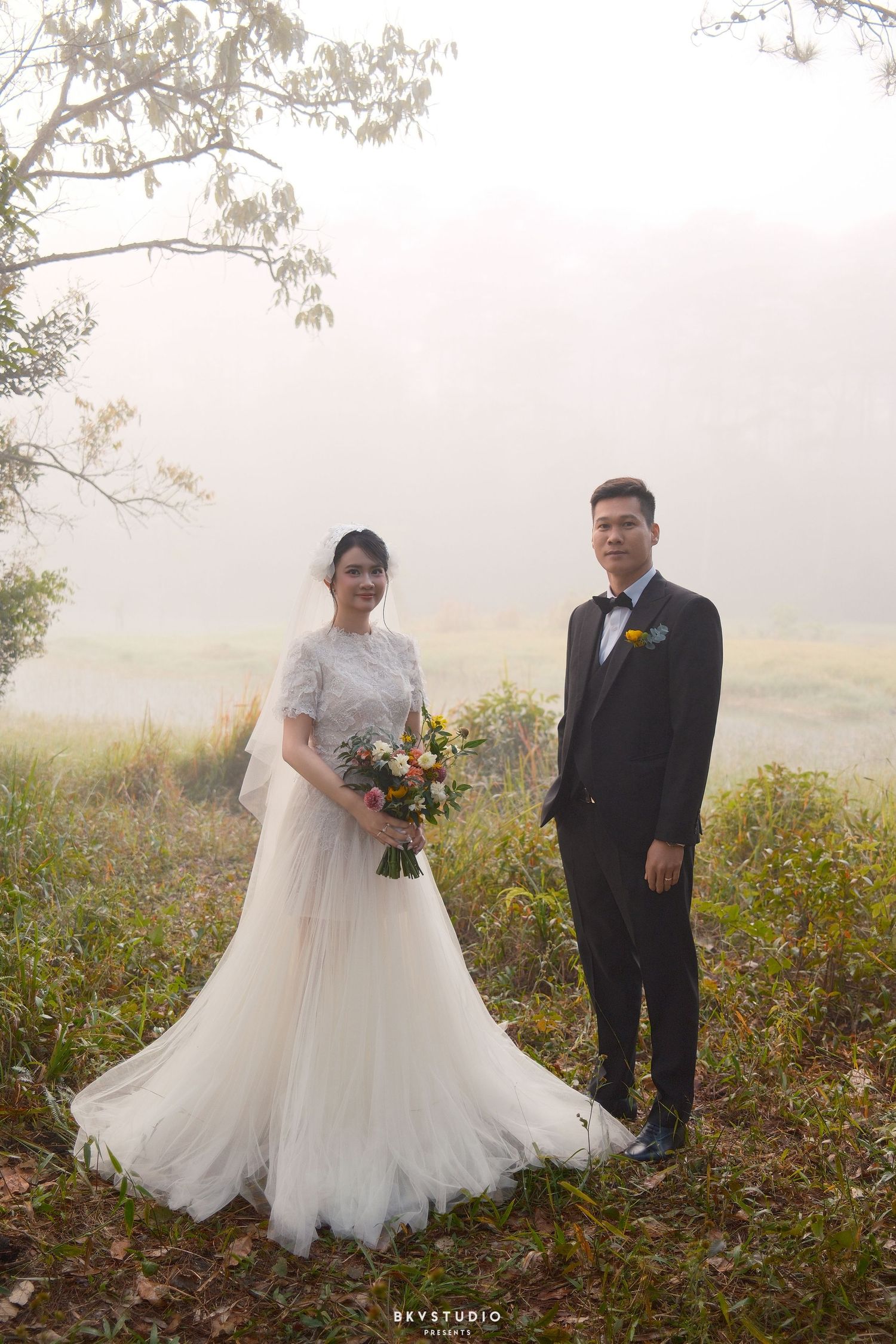 Bride in white lace gown holding bouquet stands with groom in black tuxedo in misty outdoor wedding photo.
