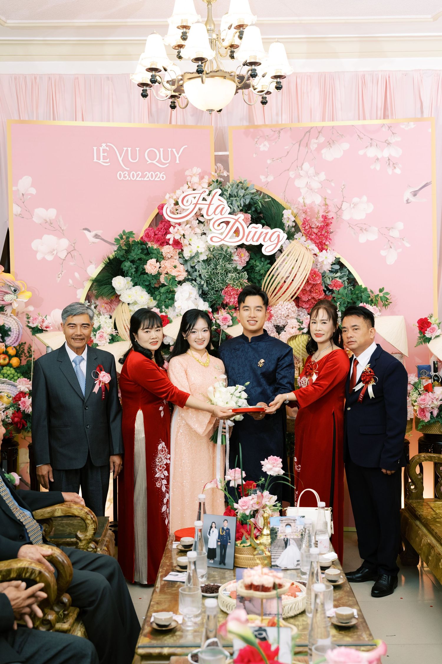 Vietnamese wedding ceremony with bride, groom, and family posing before a floral pink backdrop reading Le Vu Quy.