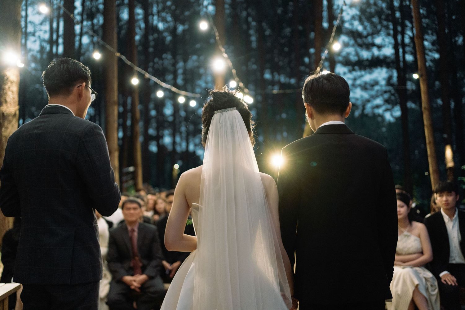 Bride and groom stand at outdoor forest wedding ceremony with string lights and seated guests.