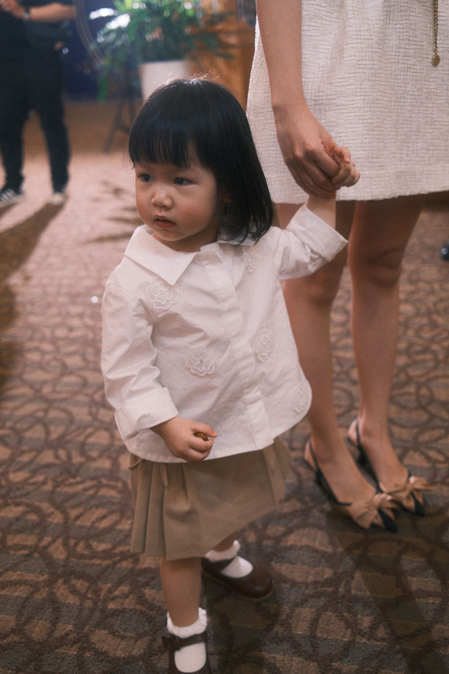 Young Asian toddler girl in white shirt and khaki skirt holds adult's hand at formal event.