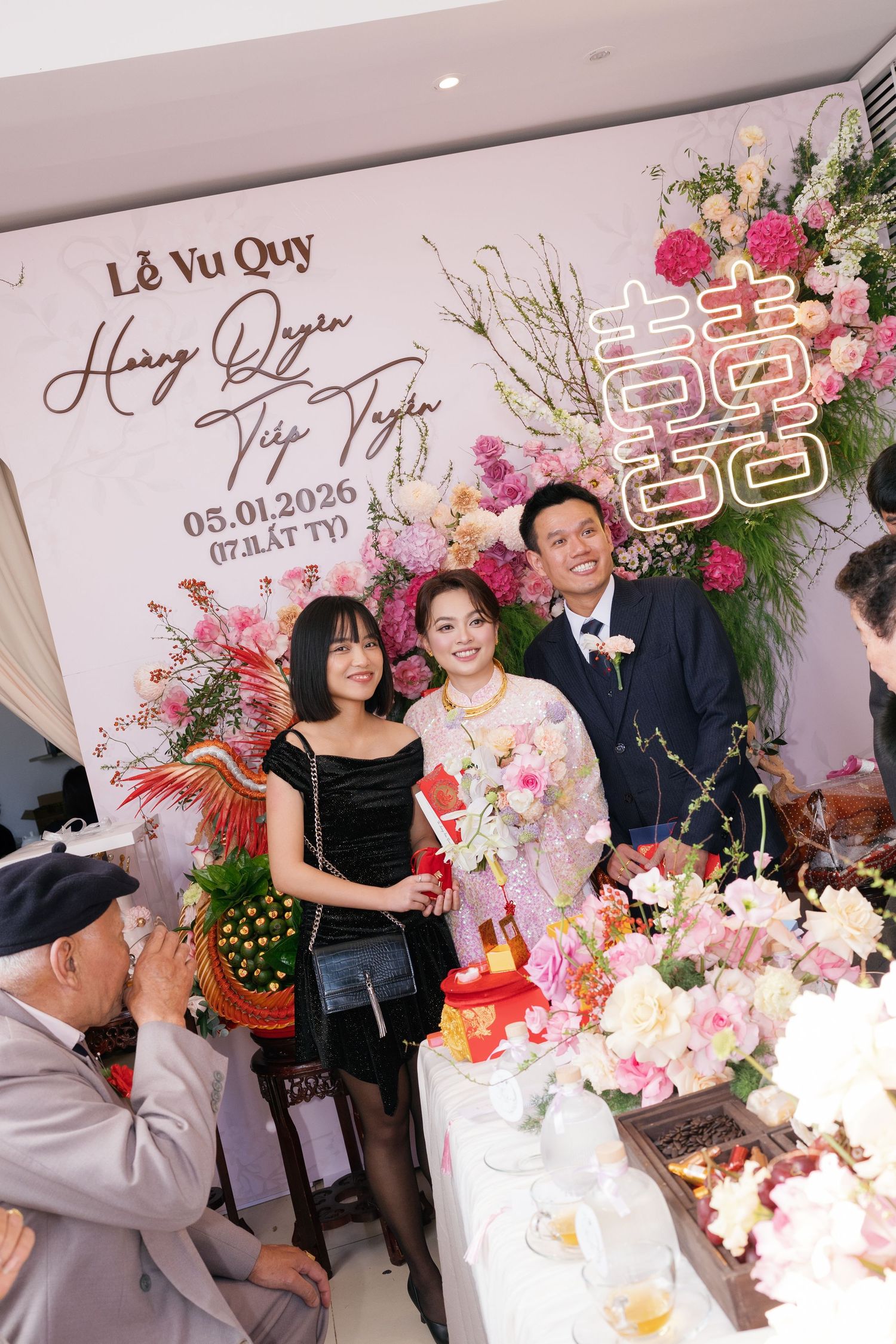 A bride in a pink ao dai poses with guests at a Vietnamese wedding reception with floral decor.