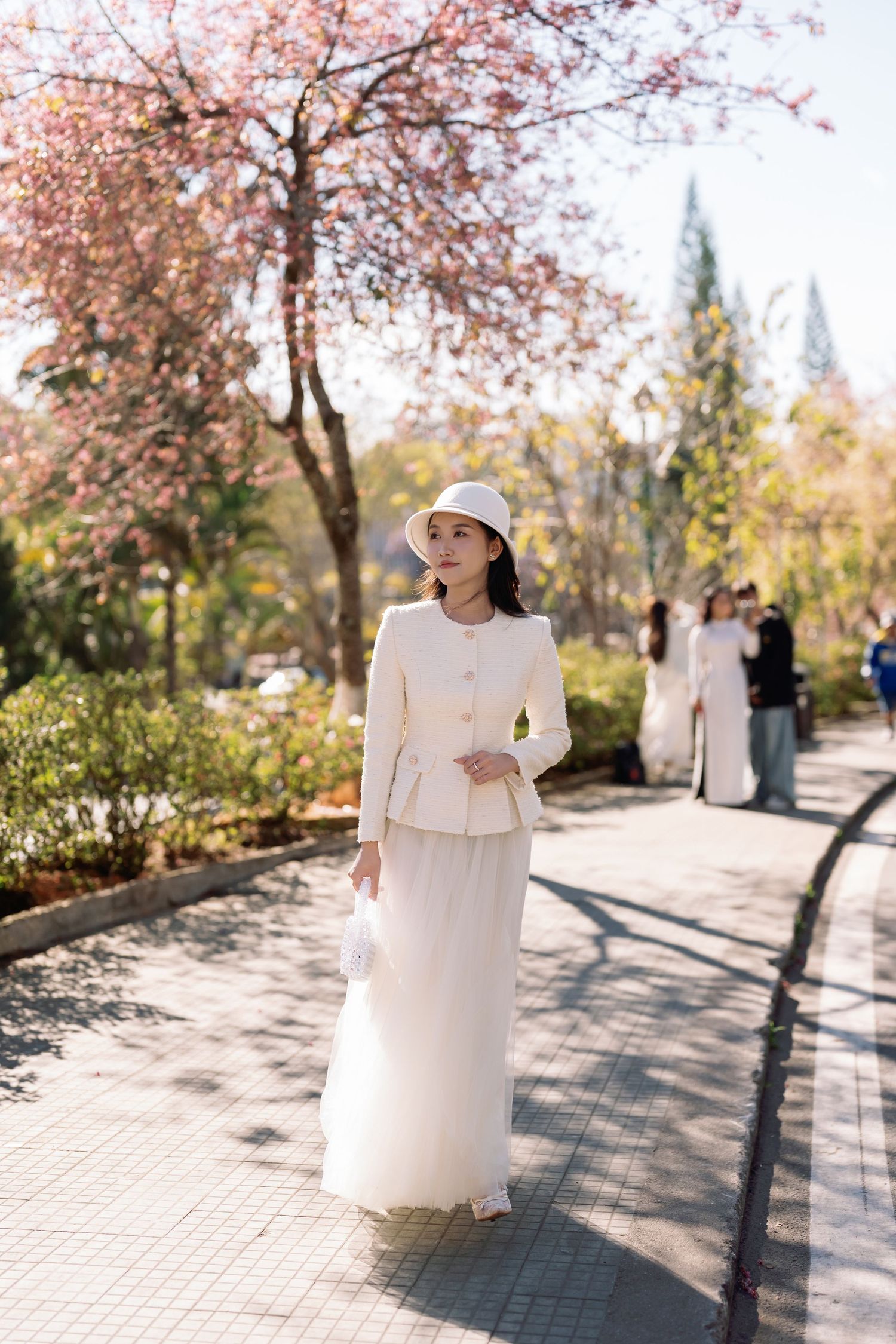 Woman in white outfit and hat strolling along a sunny park path lined with blooming pink cherry blossom trees.
