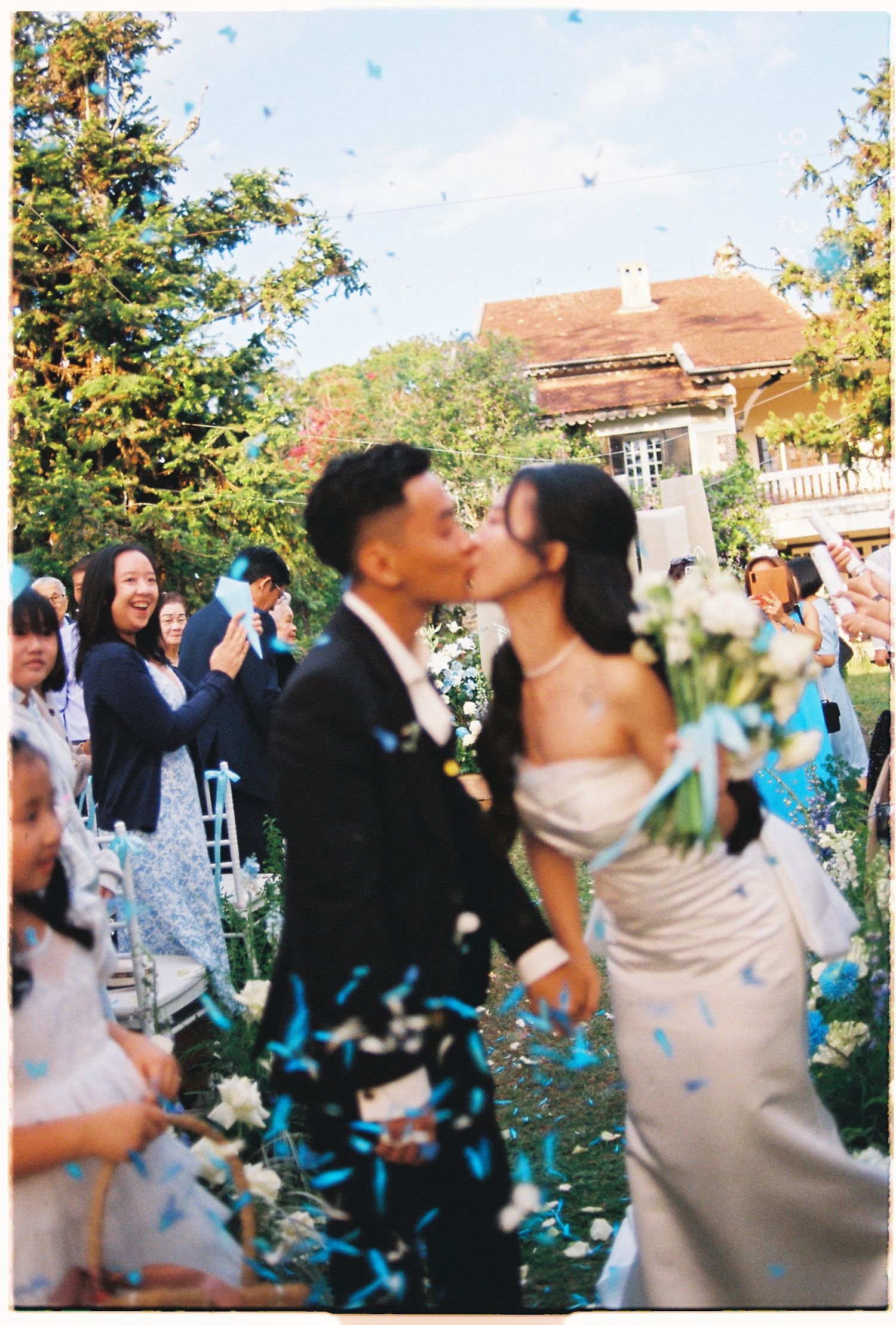 Couple sharing a kiss at outdoor wedding ceremony while guests celebrate with blue confetti falling around them.