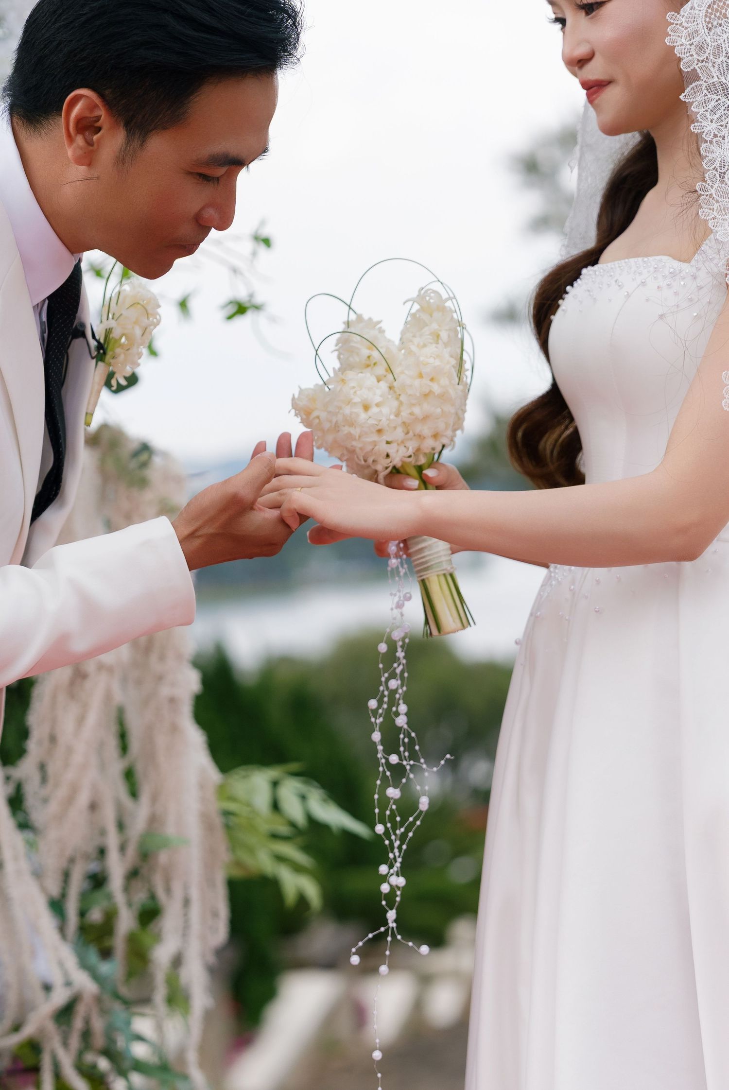 Groom in white suit kissing bride's hand while she holds elegant white floral bouquet with pearl details at outdoor wedding.