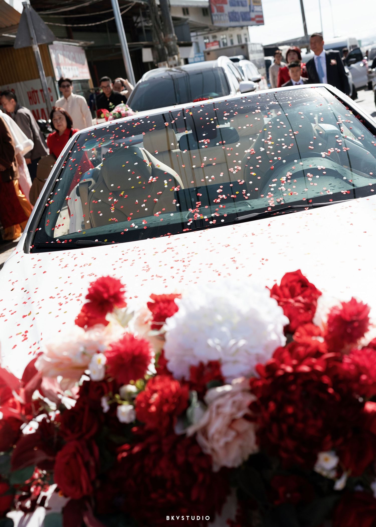 White wedding car adorned with red and white flowers and confetti petals at an outdoor wedding ceremony.
