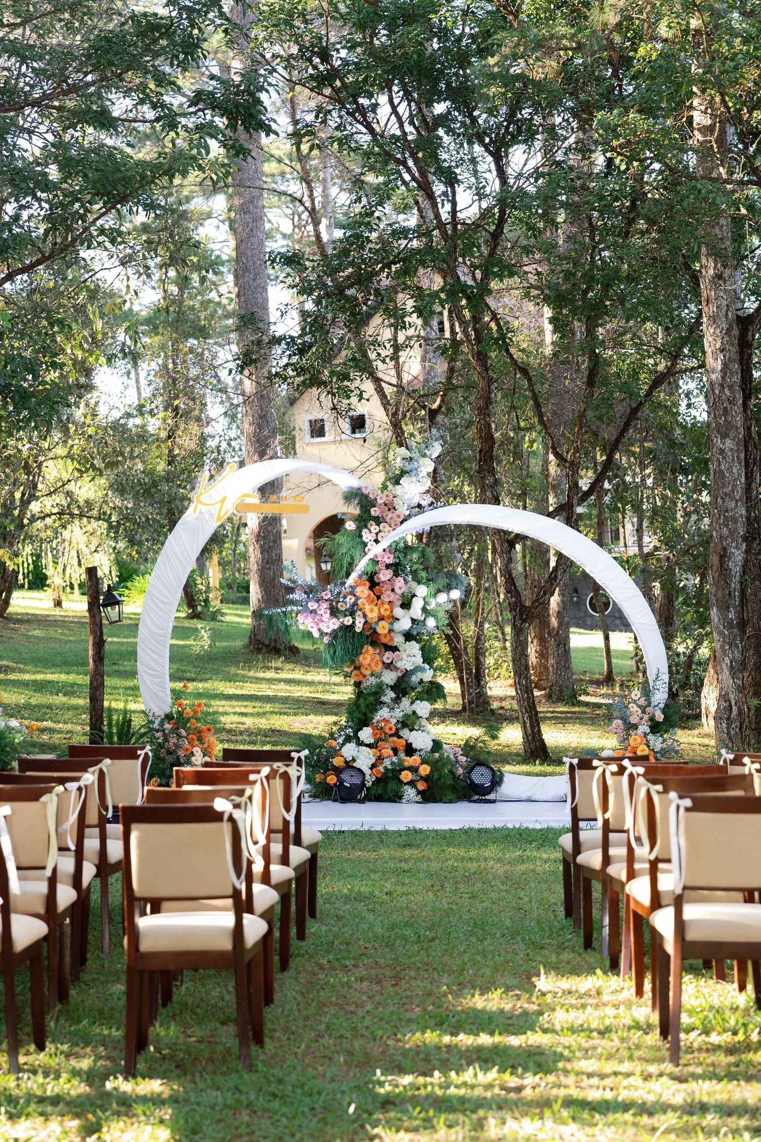 Elegant outdoor wedding ceremony setup with circular floral arch, colorful blooms, and wooden chairs on green lawn.