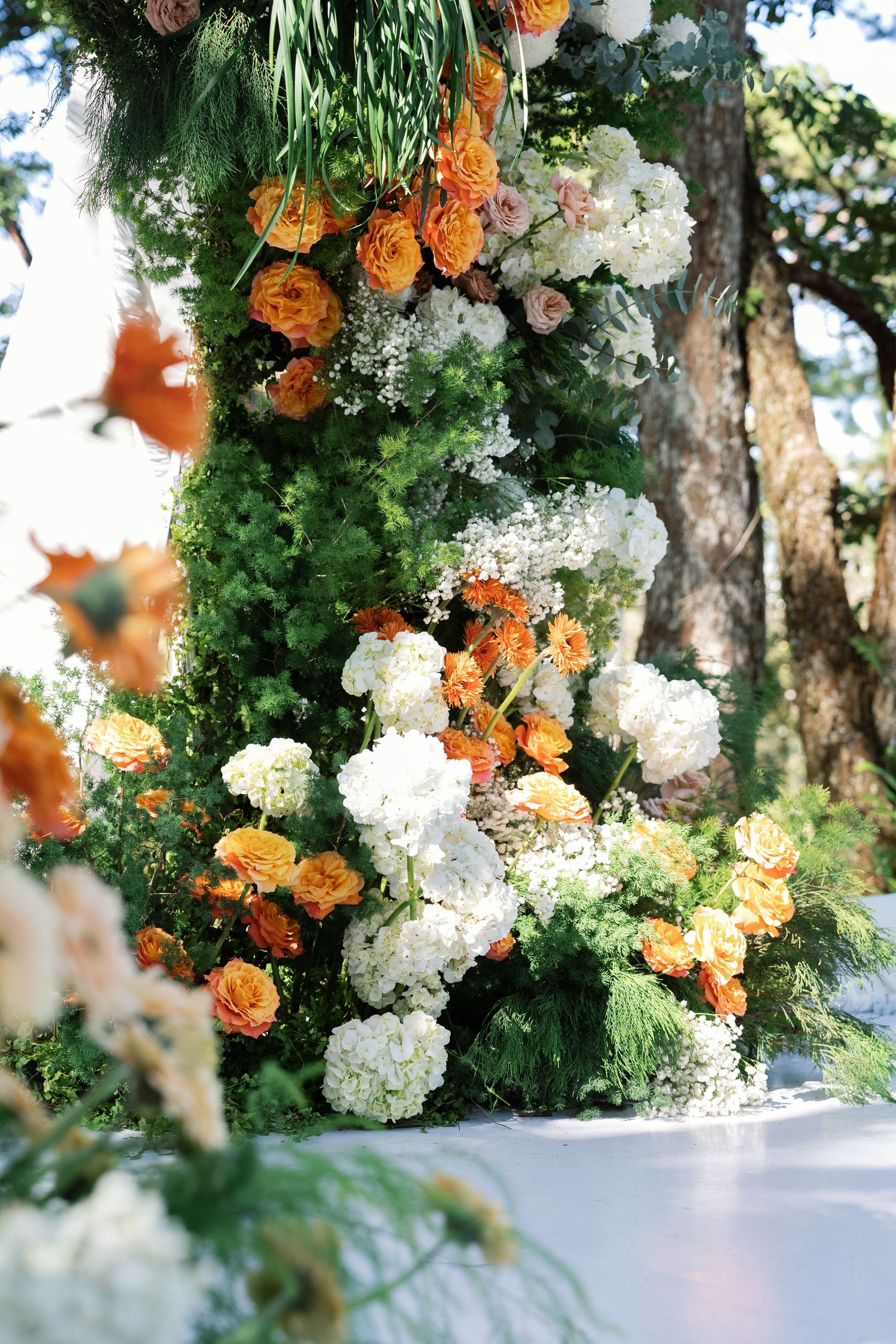 Lush wedding arch adorned with orange roses, white hydrangeas, and green foliage outdoors.