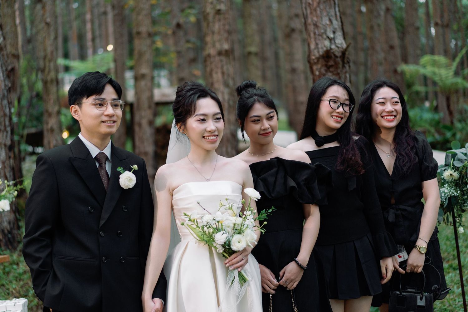 Wedding party posing in a forest setting, bride in white gown holding bouquet with groom and guests in formal attire.