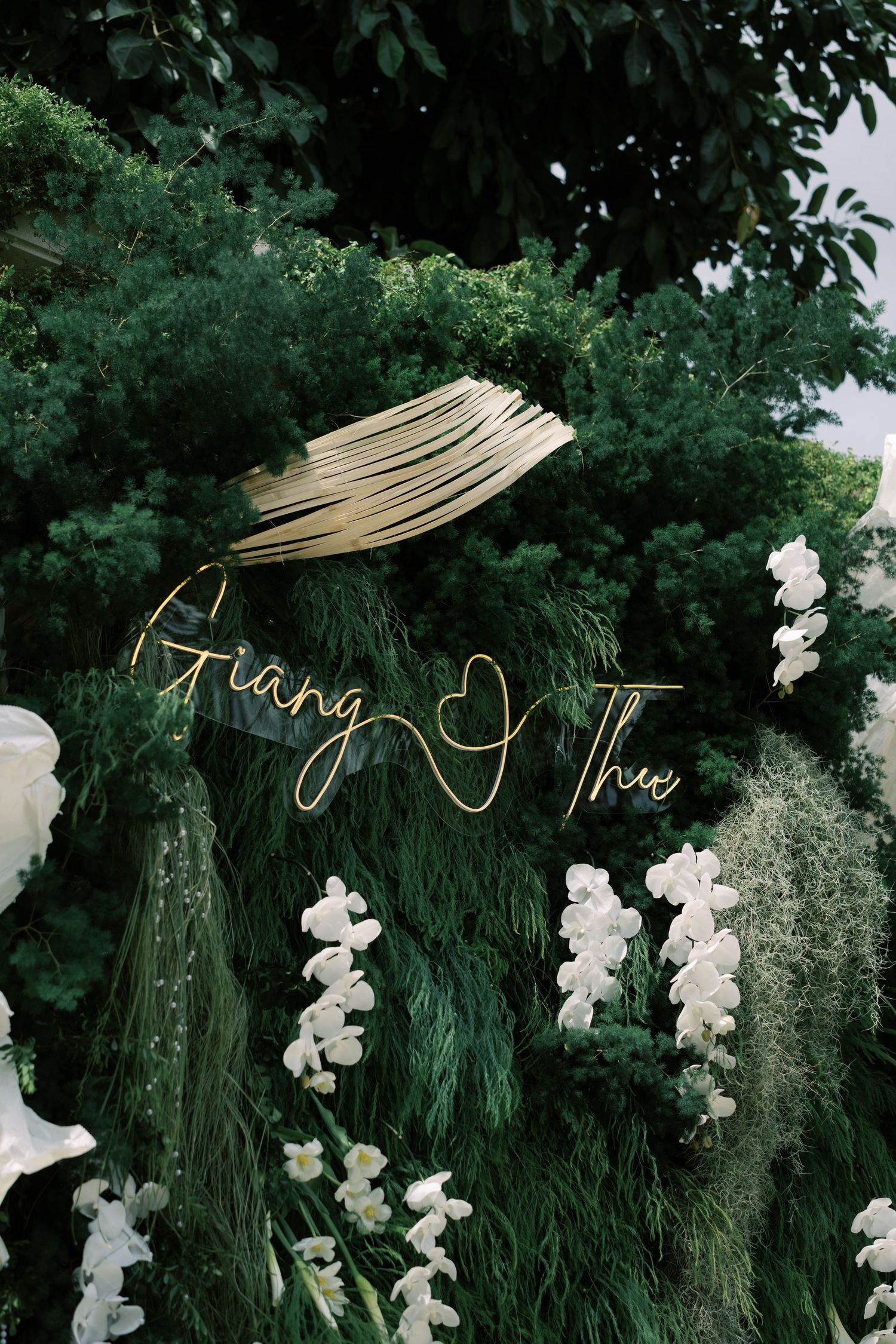 Lush green floral wedding backdrop with gold neon sign reading Giang and Thu, adorned with white orchids.