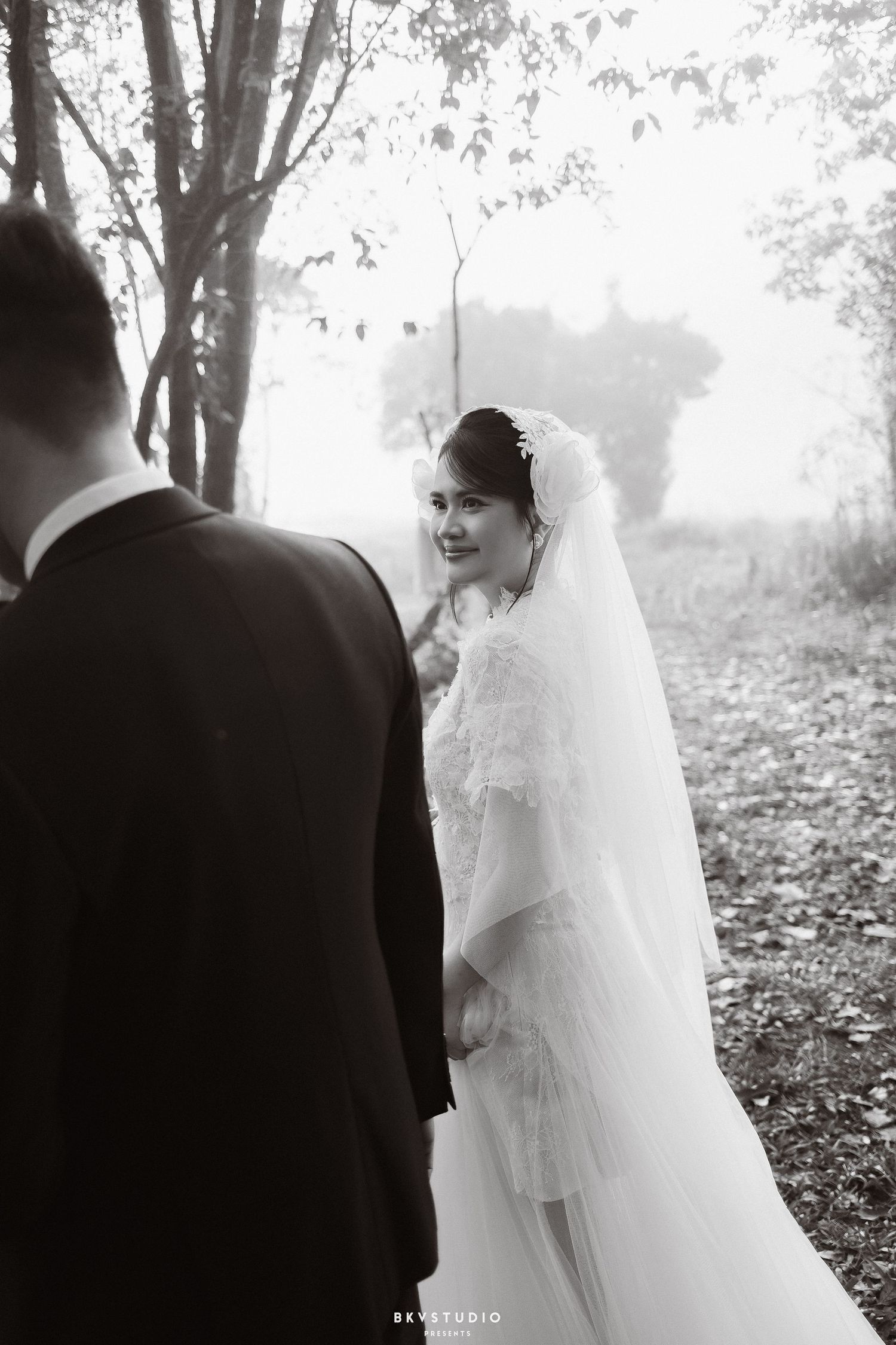 Black and white wedding photo of smiling bride in veil looking back at groom in misty forest setting.