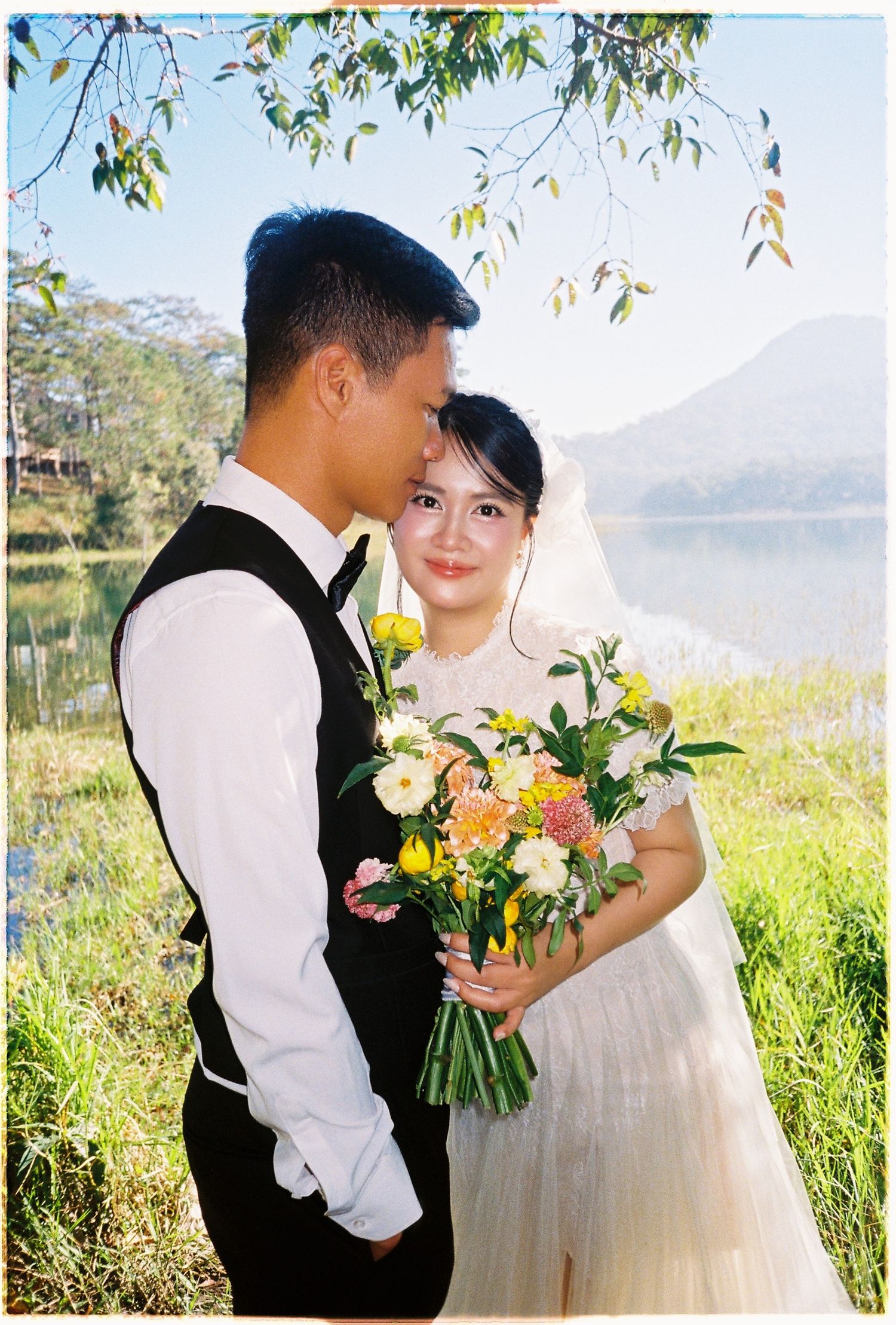 Groom tenderly kisses bride beside a sunlit lake as she holds a bright yellow and white bouquet.