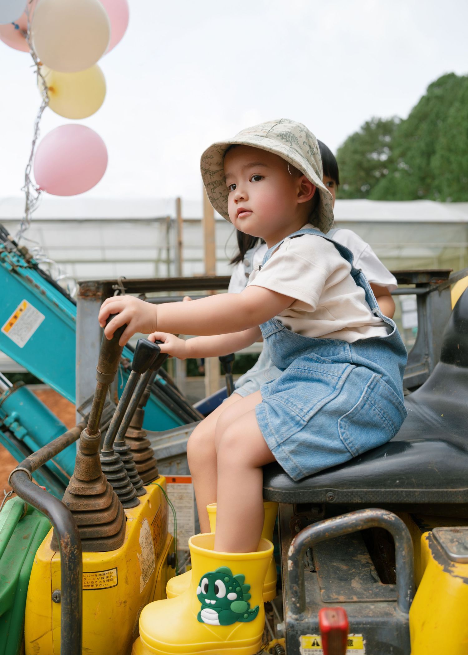 A young child in denim overalls and yellow boots sits on a tractor, wearing a bucket hat with colorful balloons nearby.