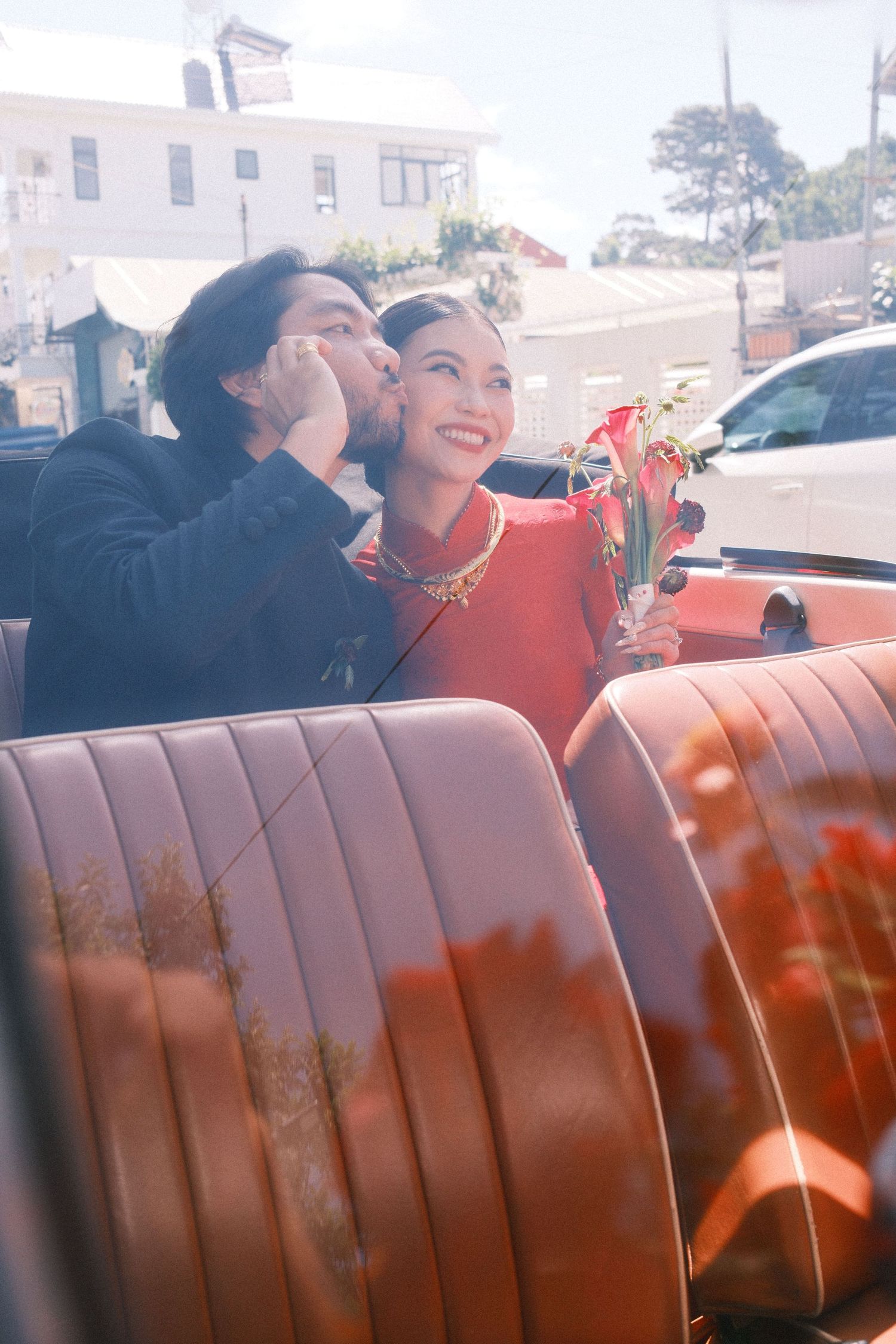 Couple in vintage convertible car, woman in red dress holding flowers, man kissing her cheek, sunny day.