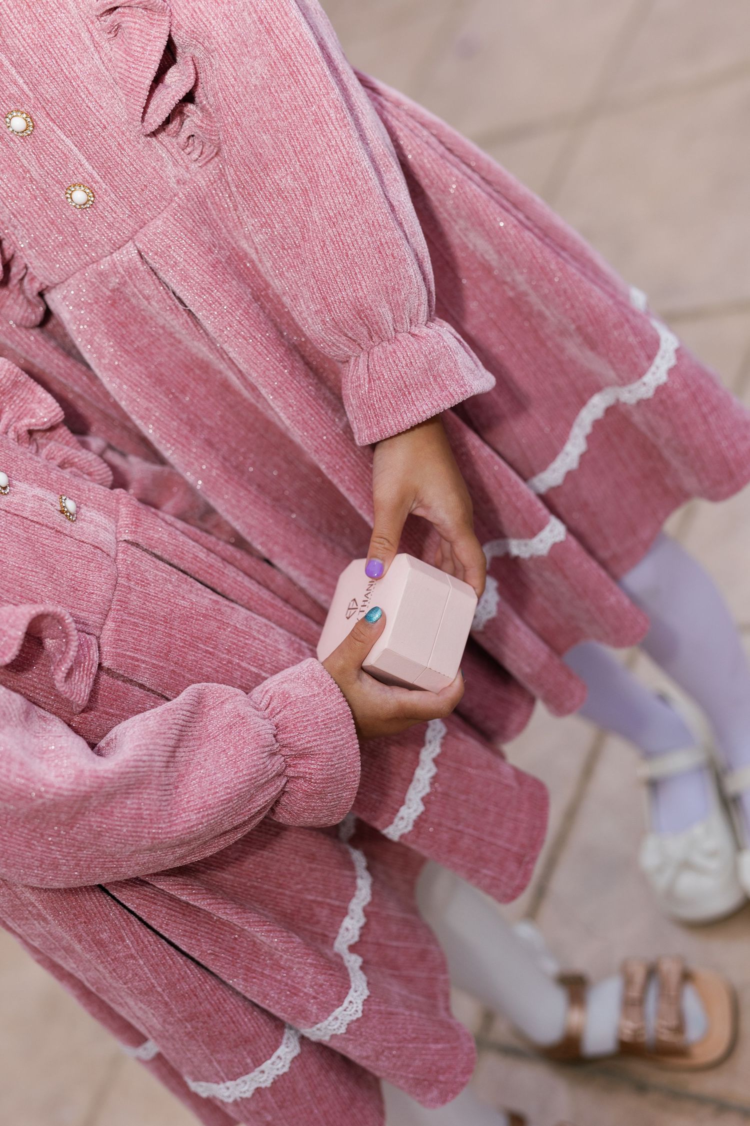 Girl in pink corduroy dress with lace trim holds a small pink box, wearing white tights and sandals.