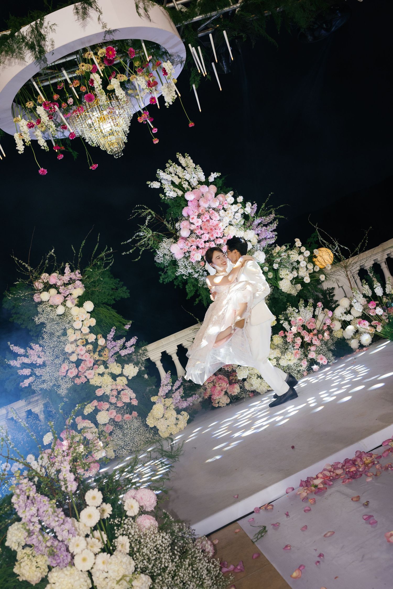 Bride and groom share a romantic kiss surrounded by lush floral arrangements at an elegant evening wedding ceremony.
