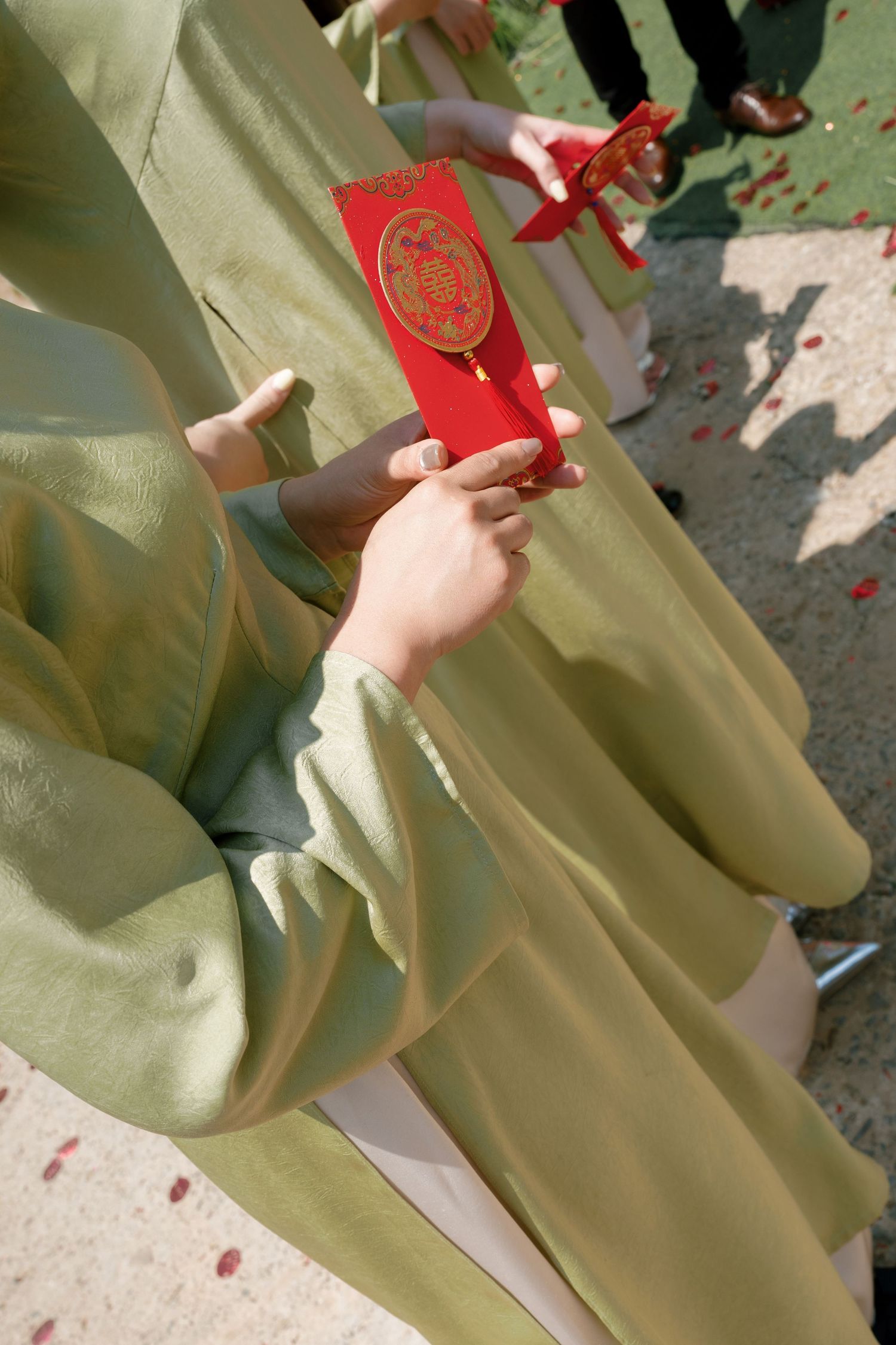 Person in green traditional Chinese attire holding a red envelope with gold double happiness symbol at a wedding ceremony.