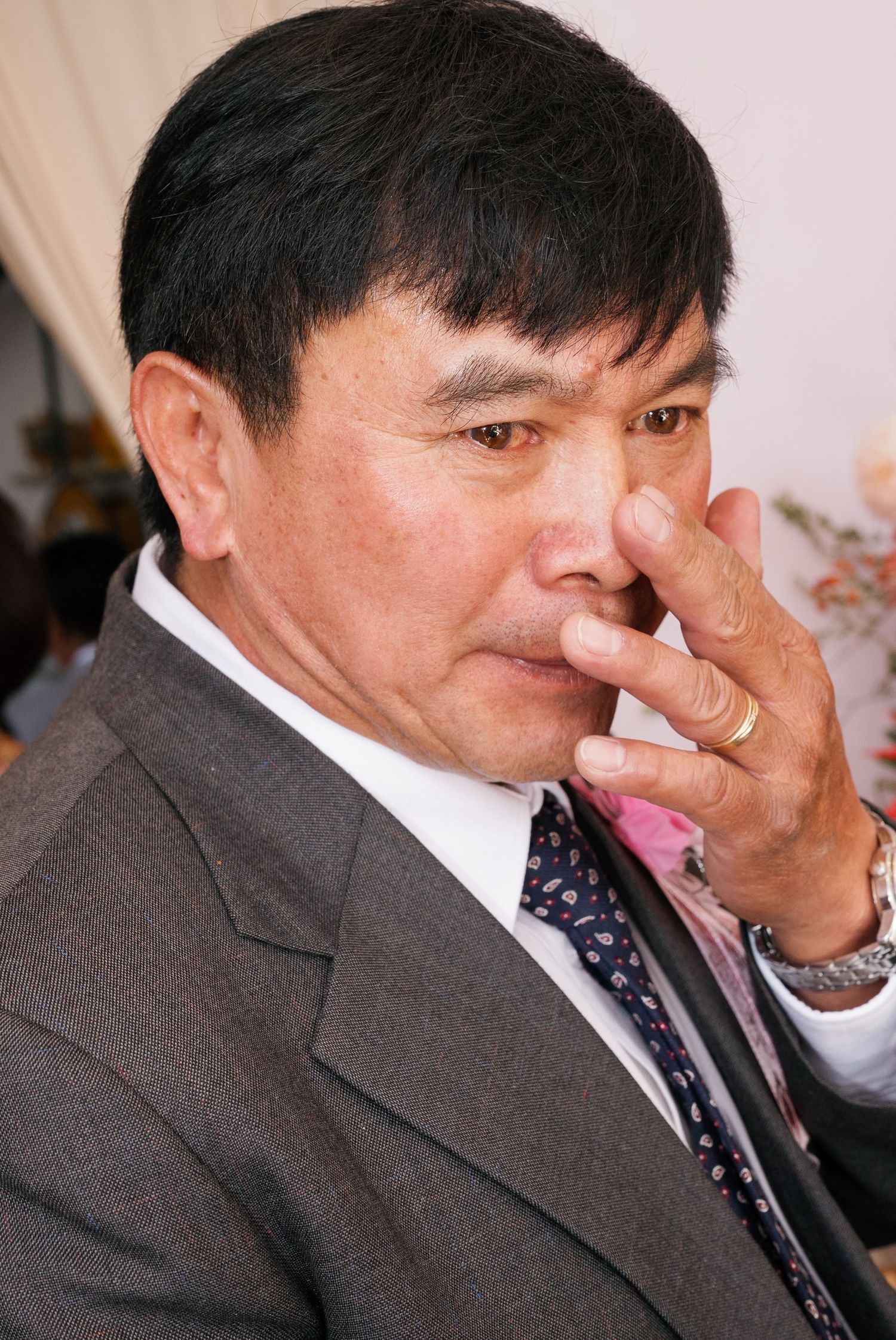 A middle-aged man in a gray suit and tie touches his face at a formal event.