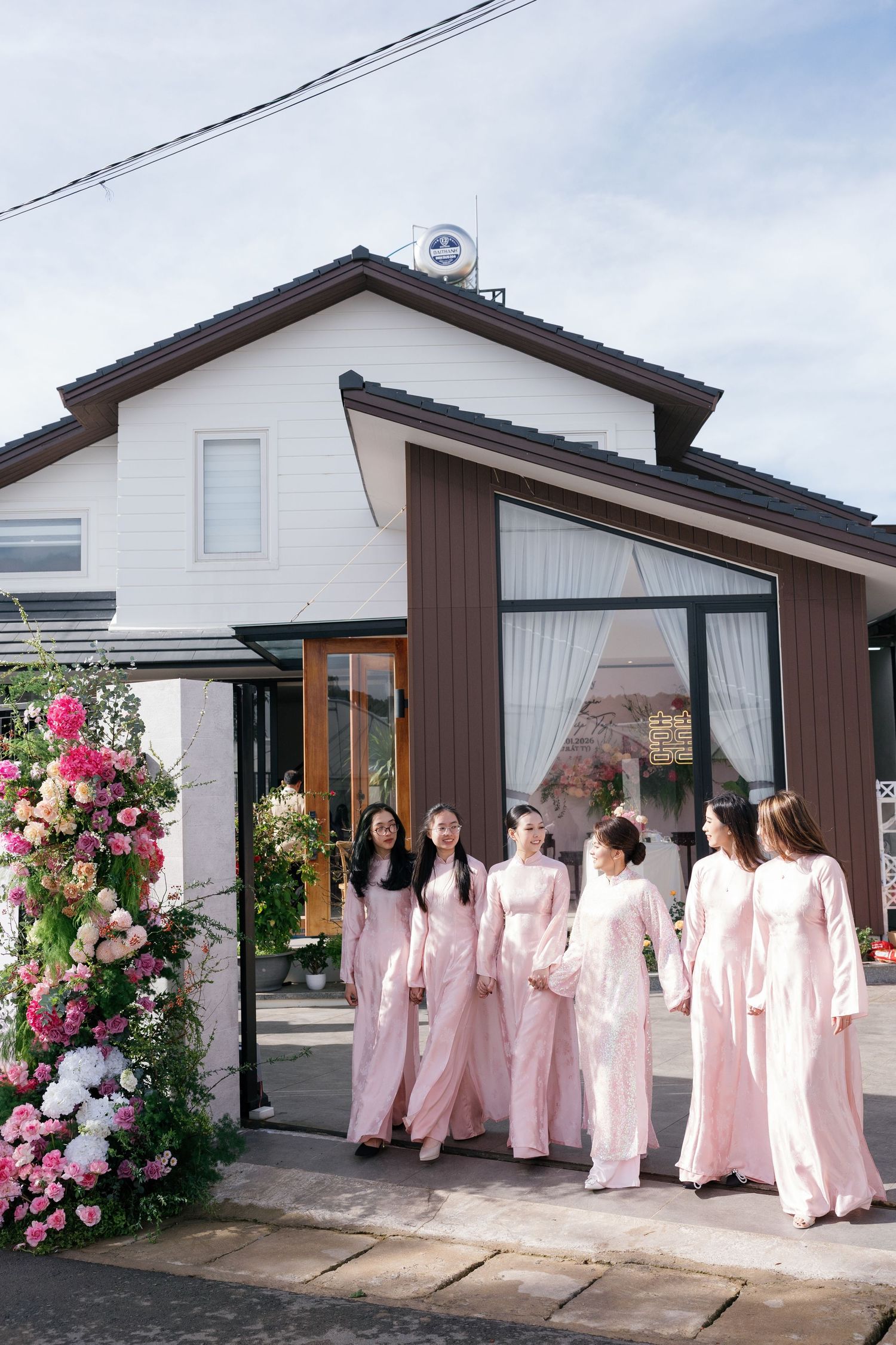 Bridesmaids in matching pink ao dai gowns walking outside a decorated house.