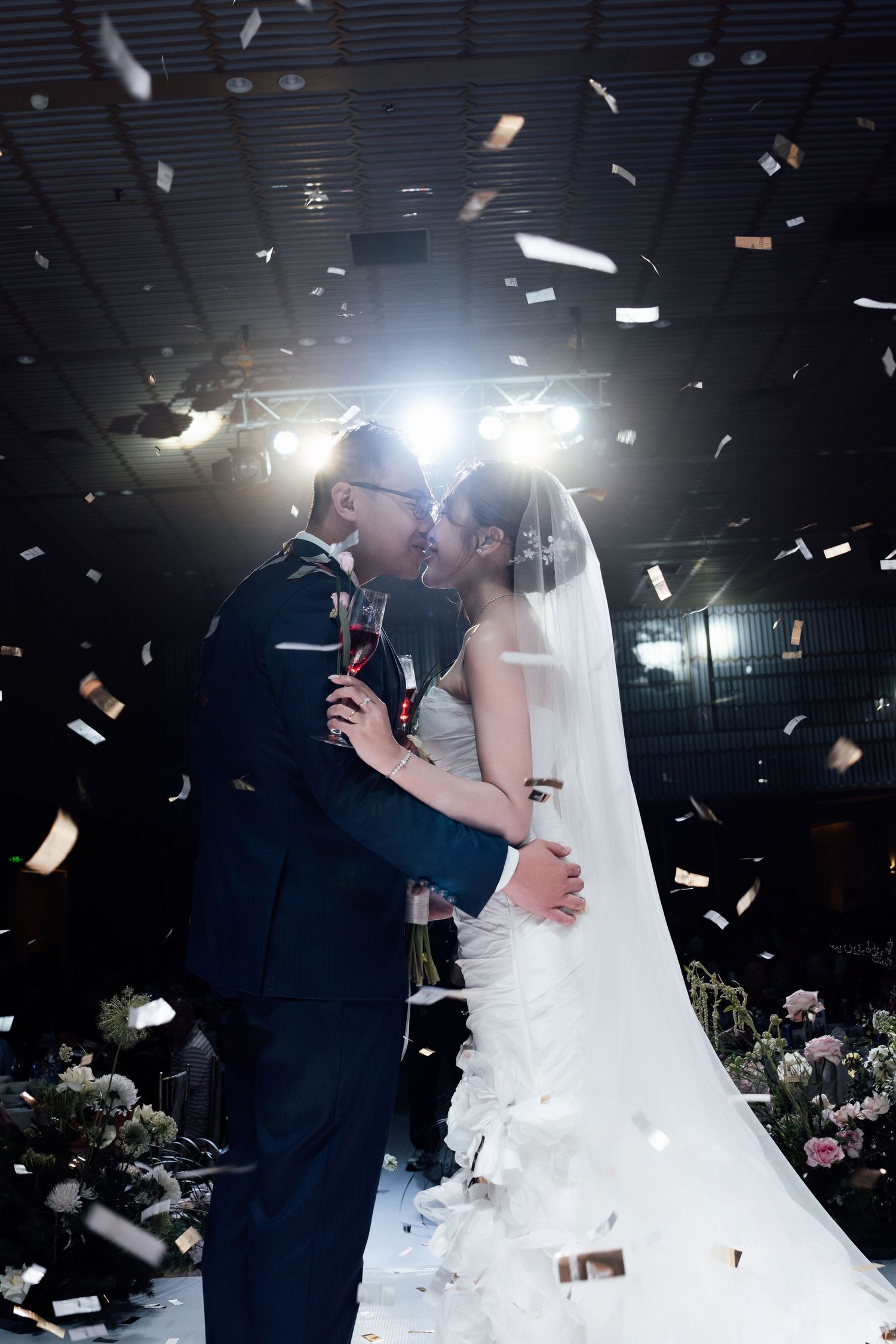 Newlyweds share first kiss surrounded by falling confetti under bright spotlight at indoor wedding ceremony.